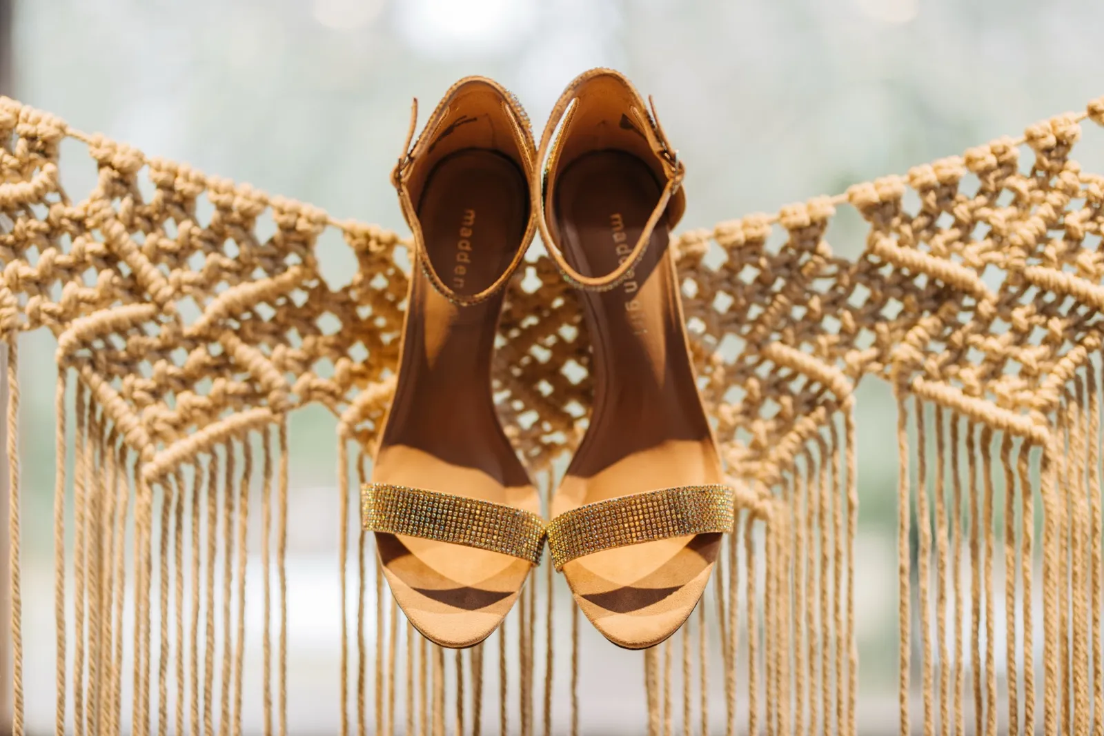 A pair of rhinestone-embellished bridal heels staged against a fringed macrame backdrop with soft window light behind. — Tim Larsen Photography, Brainerd Lakes MN