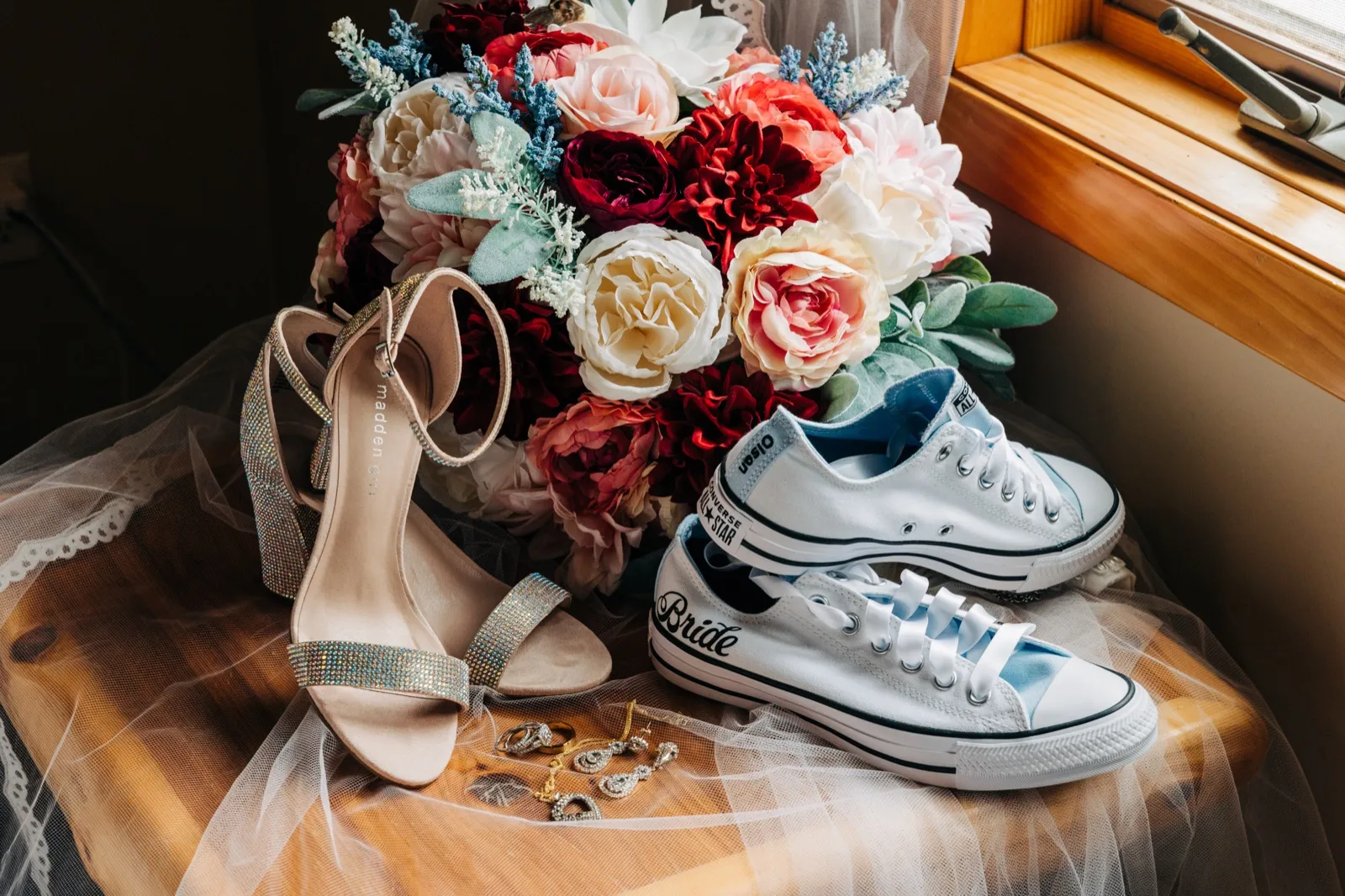 A bridal bouquet of pink, white, and burgundy blooms sits beside rhinestone heels and custom blue Converse sneakers reading 'Bride,' with earrings and rings arranged on a veil in window light. — Tim Larsen Photography, Brainerd Lakes MN