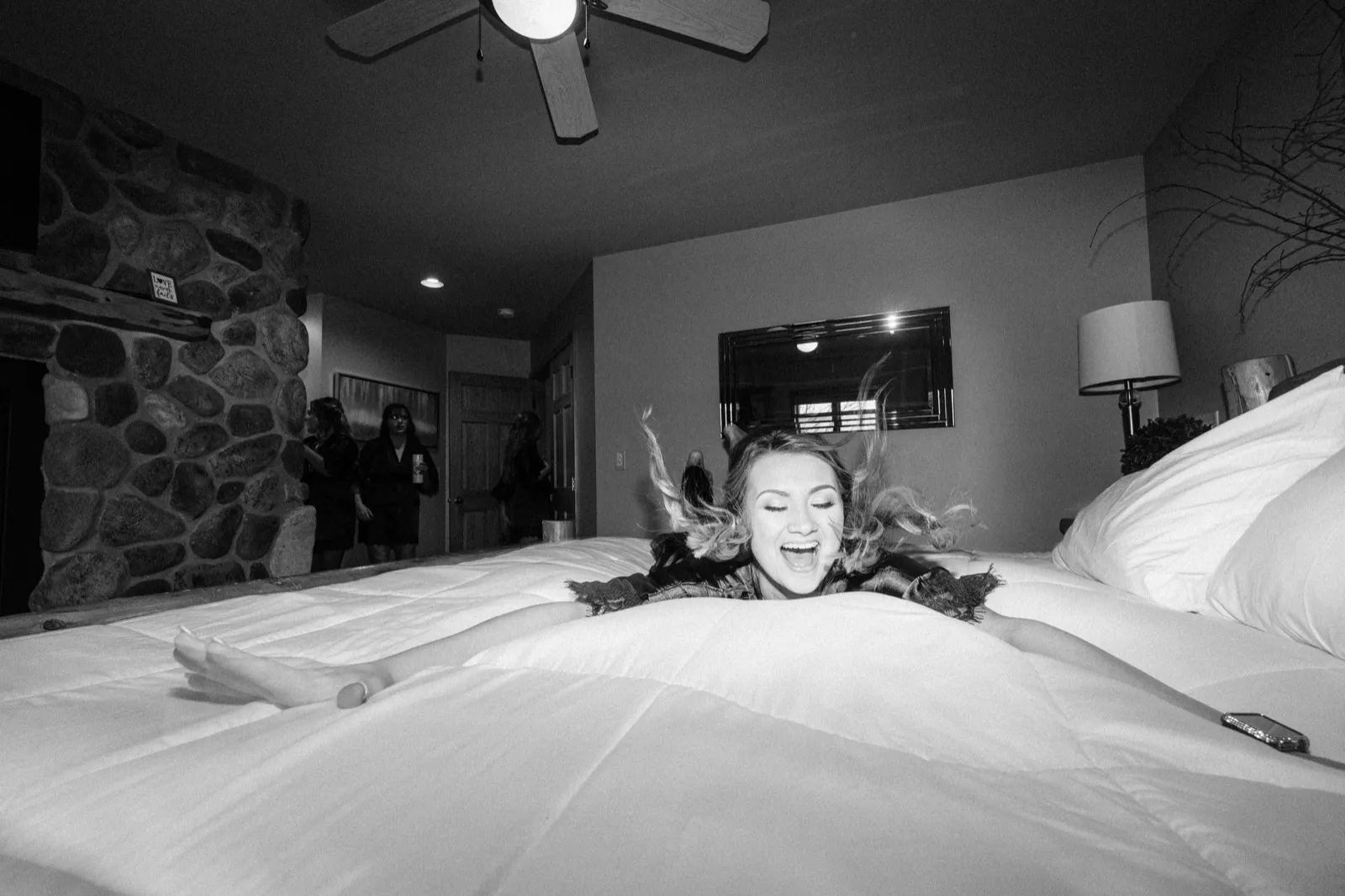 The bride laughs with her hair flying as she flops face-down onto a king-size bed in a stone-walled cabin bedroom, with bridesmaids visible in the background. — Tim Larsen Photography, Brainerd Lakes MN