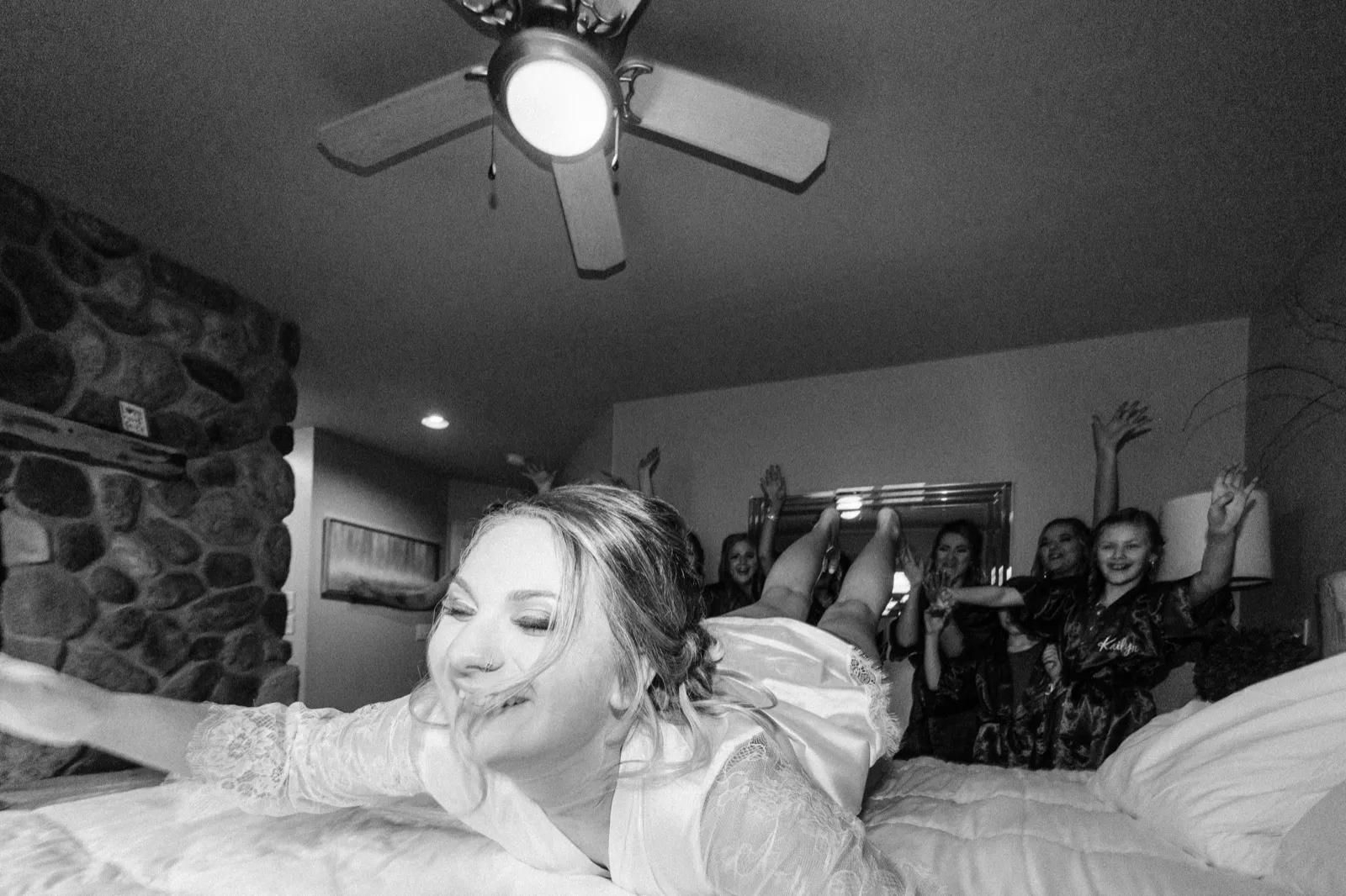 The bride lies across a bed in her robe smiling while her bridesmaids kick their feet up and cheer behind her under a ceiling fan. — Tim Larsen Photography, Brainerd Lakes MN