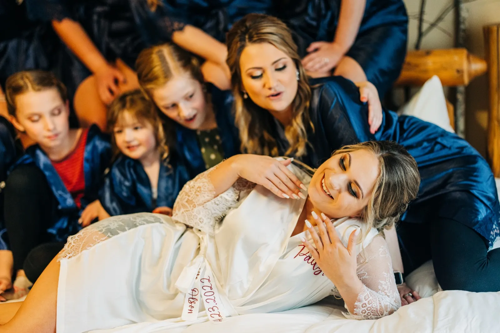 The bride reclines on a bed laughing as her bridesmaids and young flower girls in matching navy robes gather closely around her. — Tim Larsen Photography, Brainerd Lakes MN