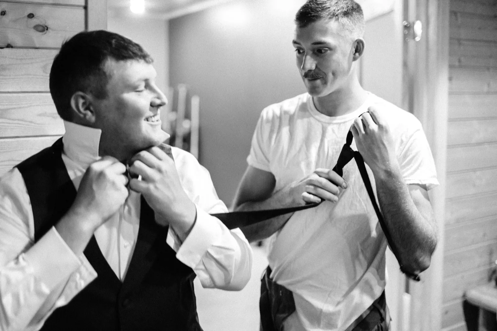A black-and-white image of a groomsman tying the groom's necktie as the groom laughs in a wood-paneled getting-ready room. — Tim Larsen Photography, Brainerd Lakes MN