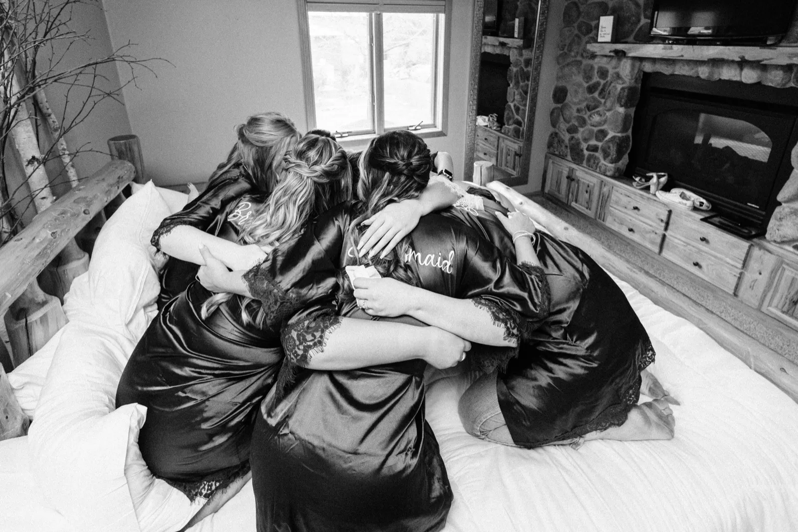 The bride and her bridesmaids in satin robes huddle together in an embrace on a bed, their backs to the camera, in a cabin room with a stone fireplace. — Tim Larsen Photography, Brainerd Lakes MN