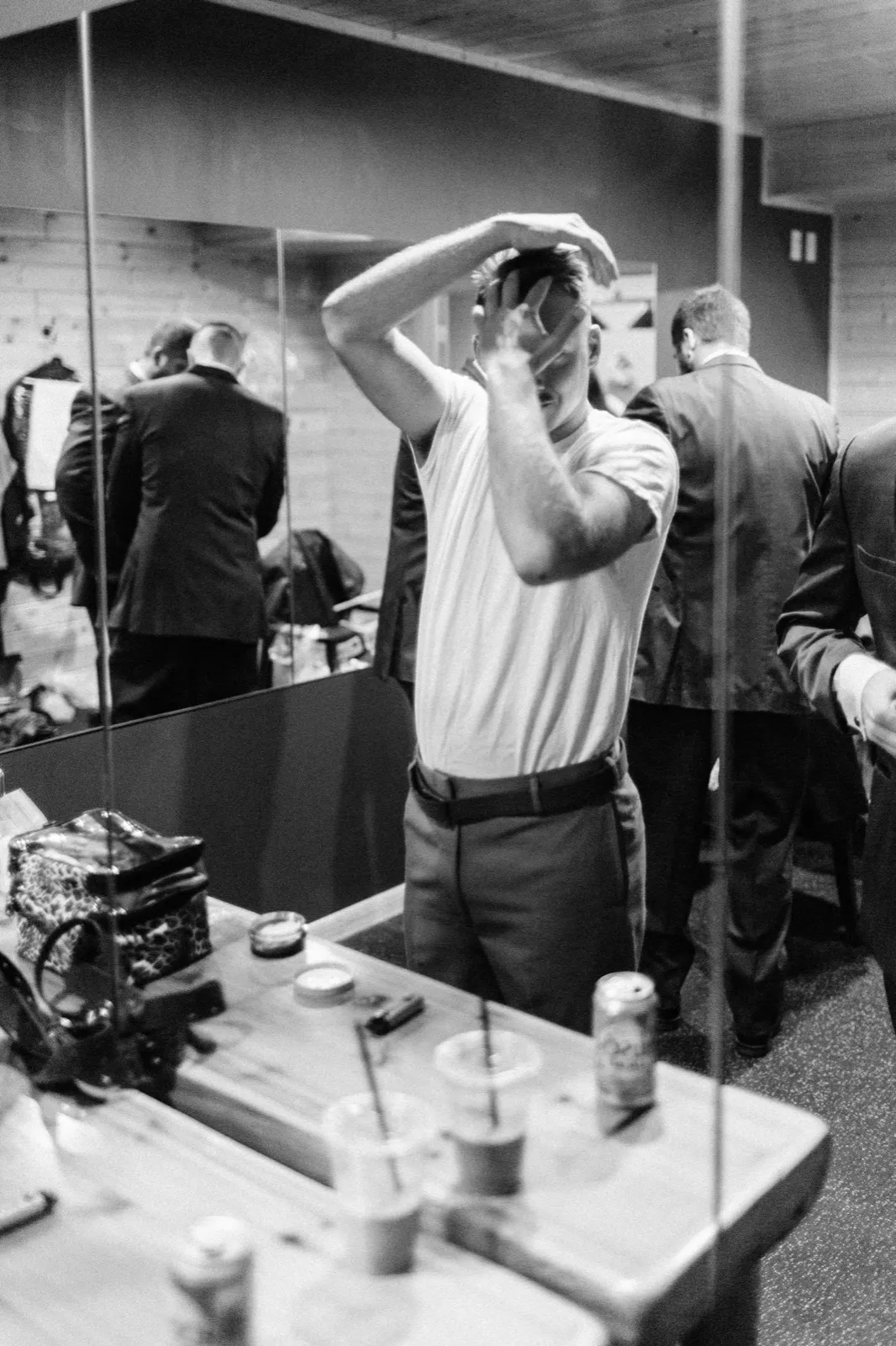 The groom in a white t-shirt fixes his hair in a mirror while groomsmen in suits stand behind him in the getting-ready room. — Tim Larsen Photography, Brainerd Lakes MN
