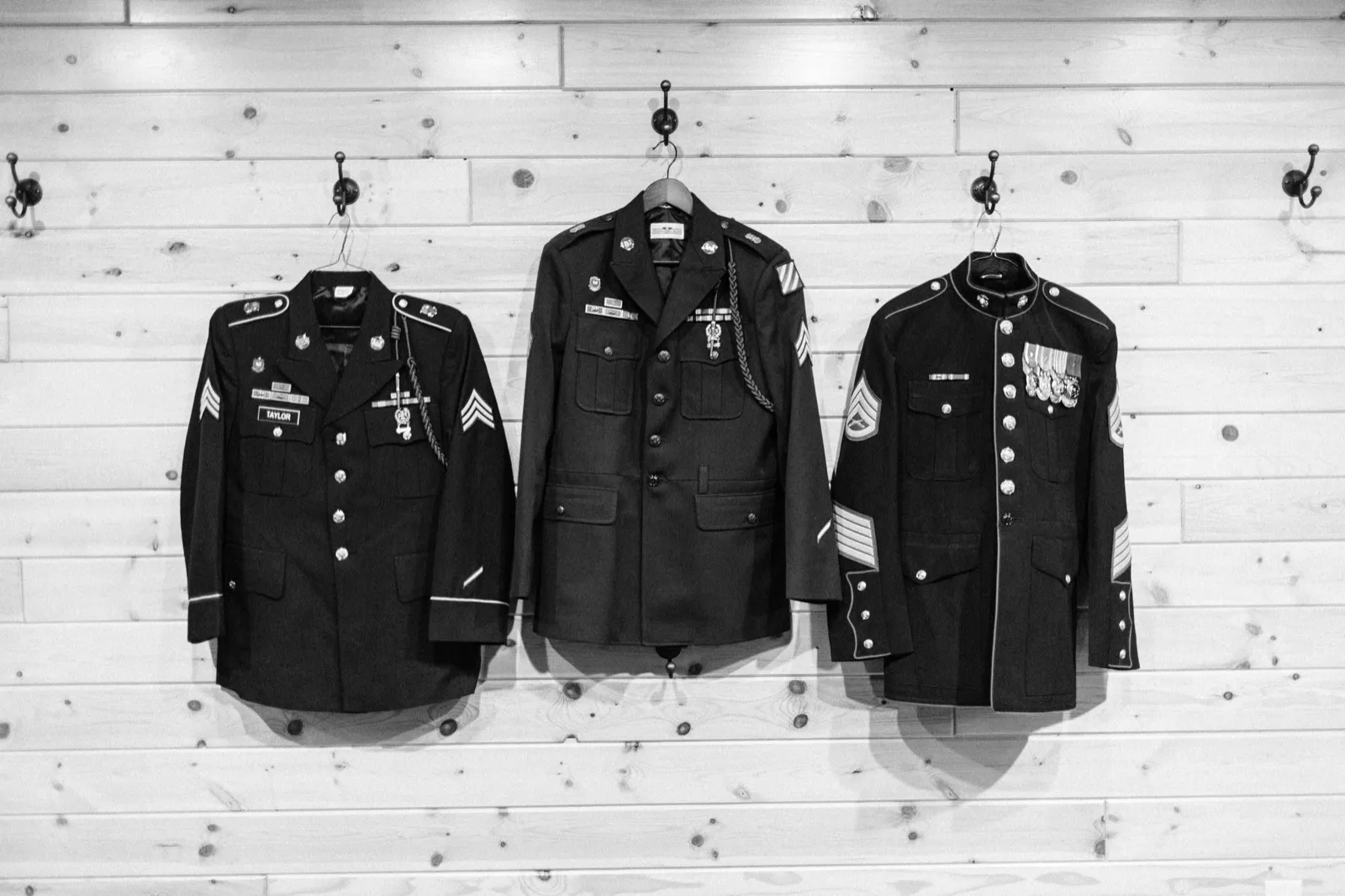 Three military dress uniforms with medals and rank insignia hang side by side on hooks against a pine plank wall, photographed in black and white. — Tim Larsen Photography, Brainerd Lakes MN