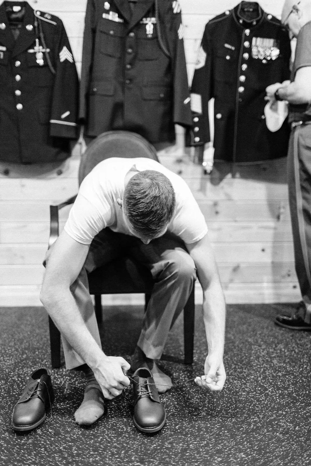 The groom sits in a t-shirt and dress pants tying his shoes beneath a wall of hung military dress uniforms heavy with medals. — Tim Larsen Photography, Brainerd Lakes MN