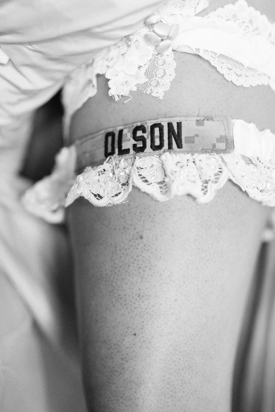 A black-and-white close-up of a lace garter on the bride's leg, embroidered with the name OLSON on a military uniform name tape. — Tim Larsen Photography, Brainerd Lakes MN