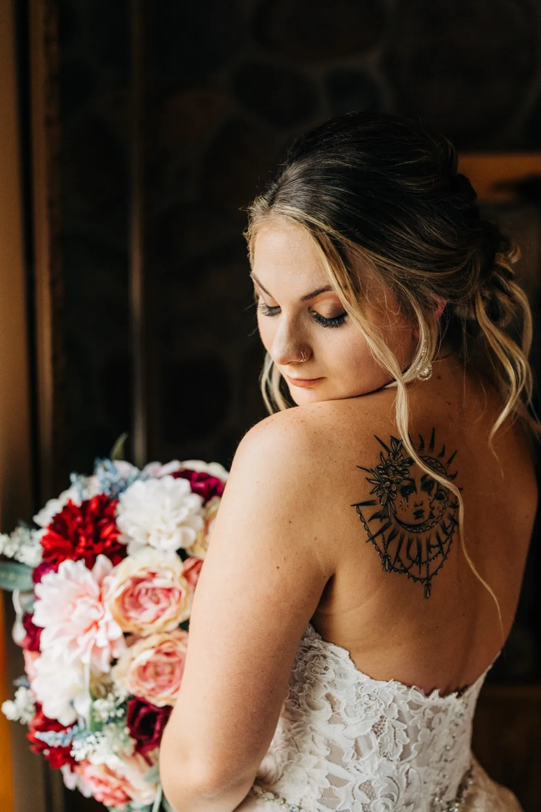 The bride looks downward over her shoulder in soft window light, revealing an ornate sun-and-moon tattoo on her back as she holds a pink and red bouquet. — Tim Larsen Photography, Brainerd Lakes MN