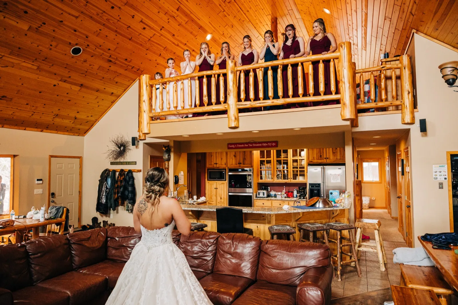 The bride stands in her gown in a cabin great room as her bridesmaids and a flower girl react with hands to their faces from a wooden loft balcony above. — Tim Larsen Photography, Brainerd Lakes MN
