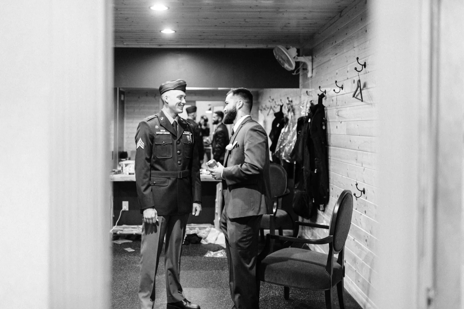 Black-and-white image viewed through a doorway showing a soldier in dress uniform and cap laughing with the groom in a dark suit, both standing in a wood-paneled hallway lined with coat hooks. — Tim Larsen Photography, Brainerd Lakes MN