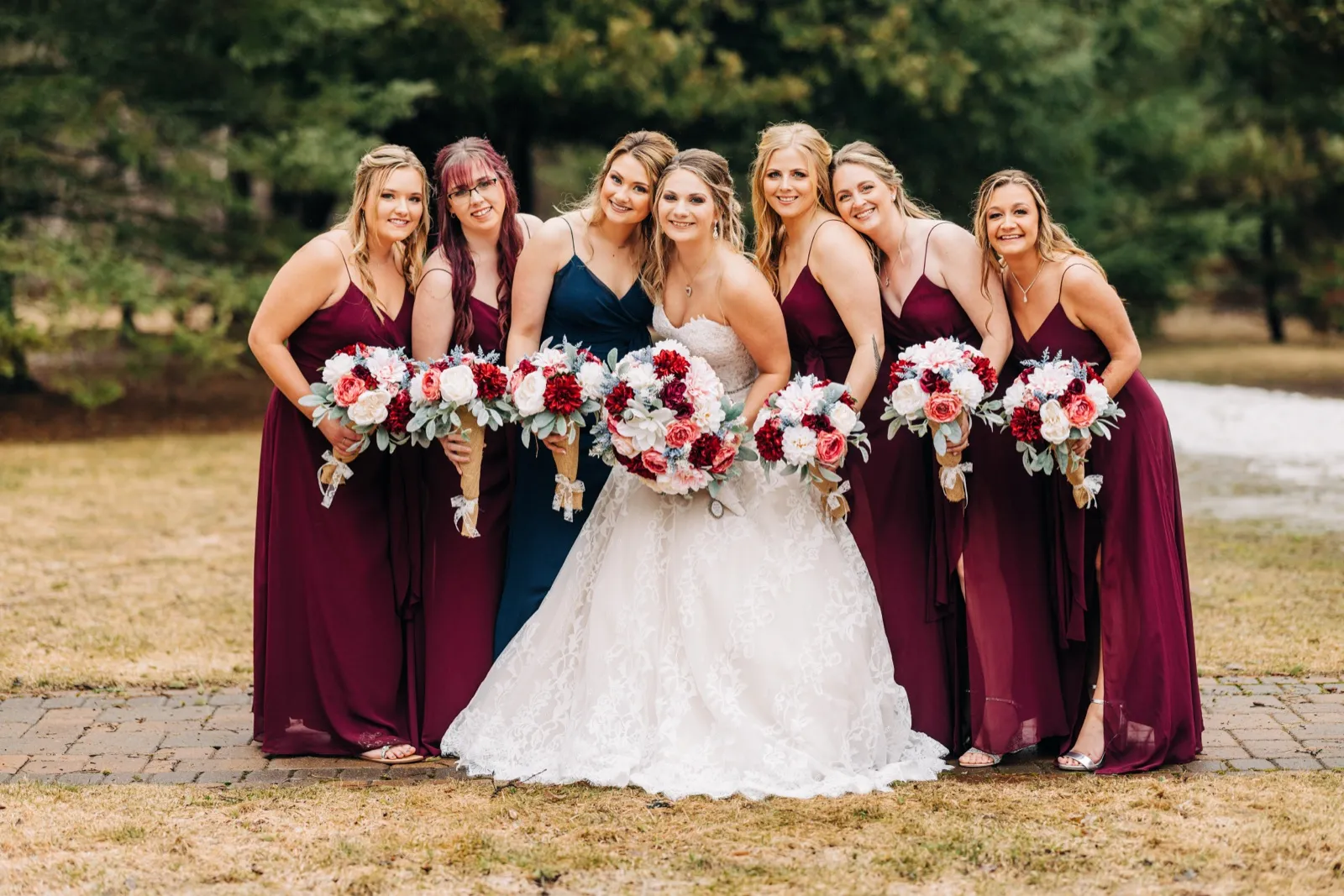 The bride and her six bridesmaids in burgundy gowns lean toward each other, holding fanned-out bouquets of pink and red florals on a wooded lawn. — Tim Larsen Photography, Brainerd Lakes MN