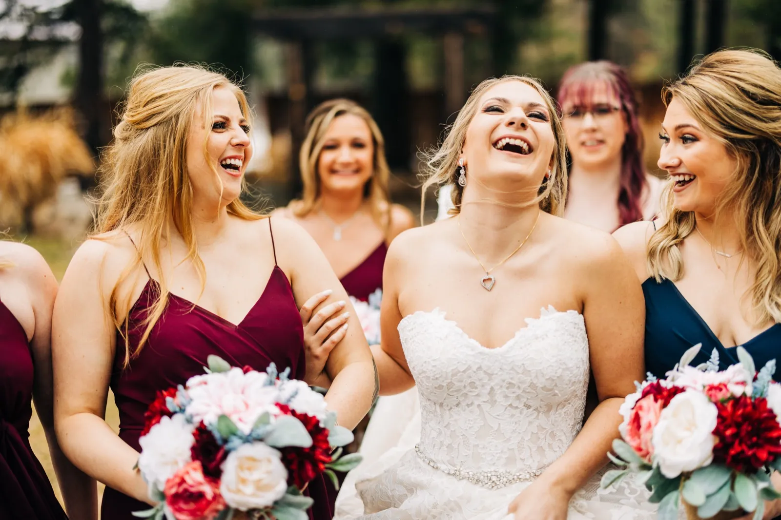The bride throws her head back laughing as her bridesmaids in burgundy and navy dresses laugh along beside her, holding red and blush bouquets. — Tim Larsen Photography, Brainerd Lakes MN
