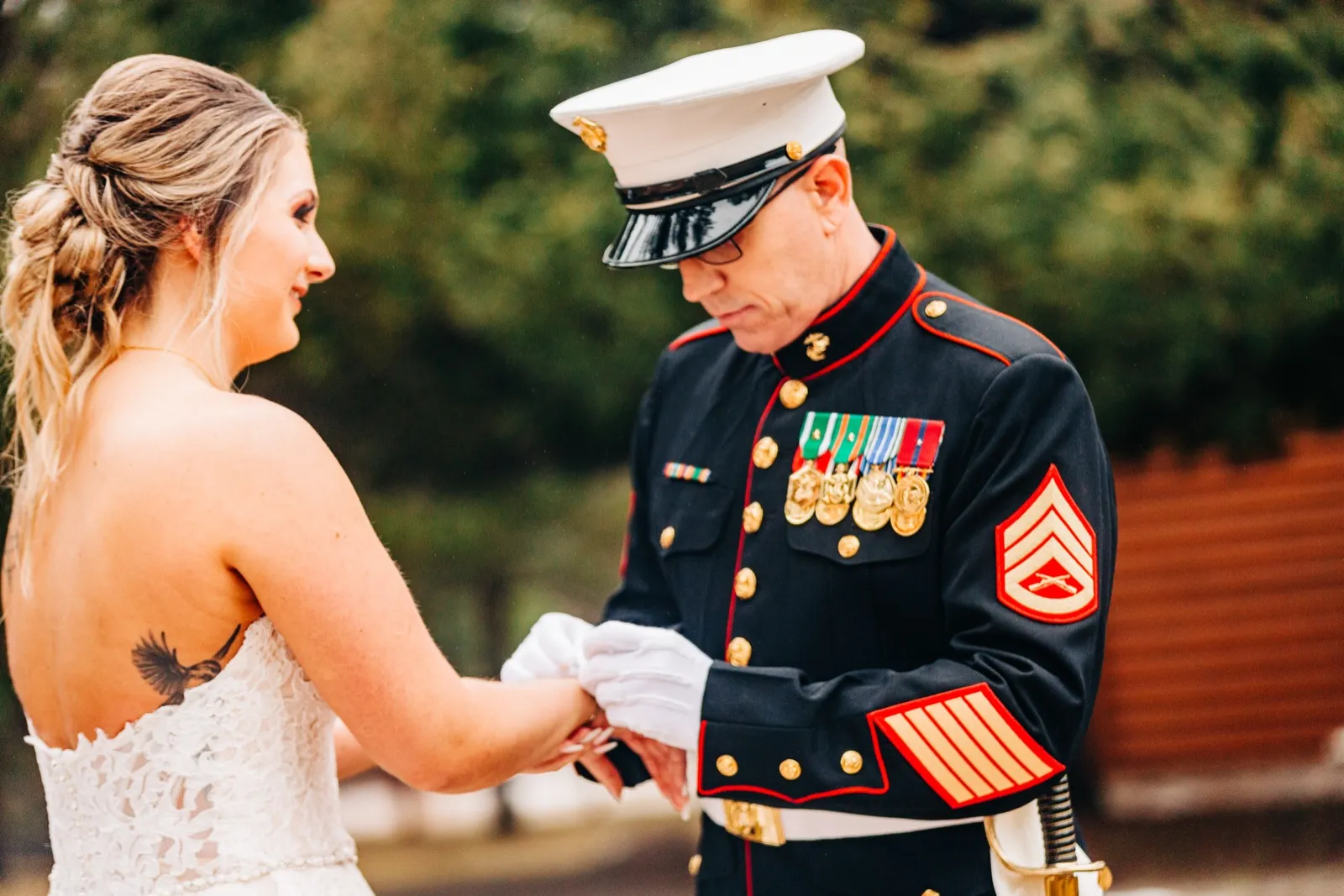 A bride in a strapless lace gown holds the white-gloved hands of her groom, who wears a Marine Corps dress blue uniform with medals and a peaked cap, his head bowed toward their joined hands. — Tim Larsen Photography, Brainerd Lakes MN