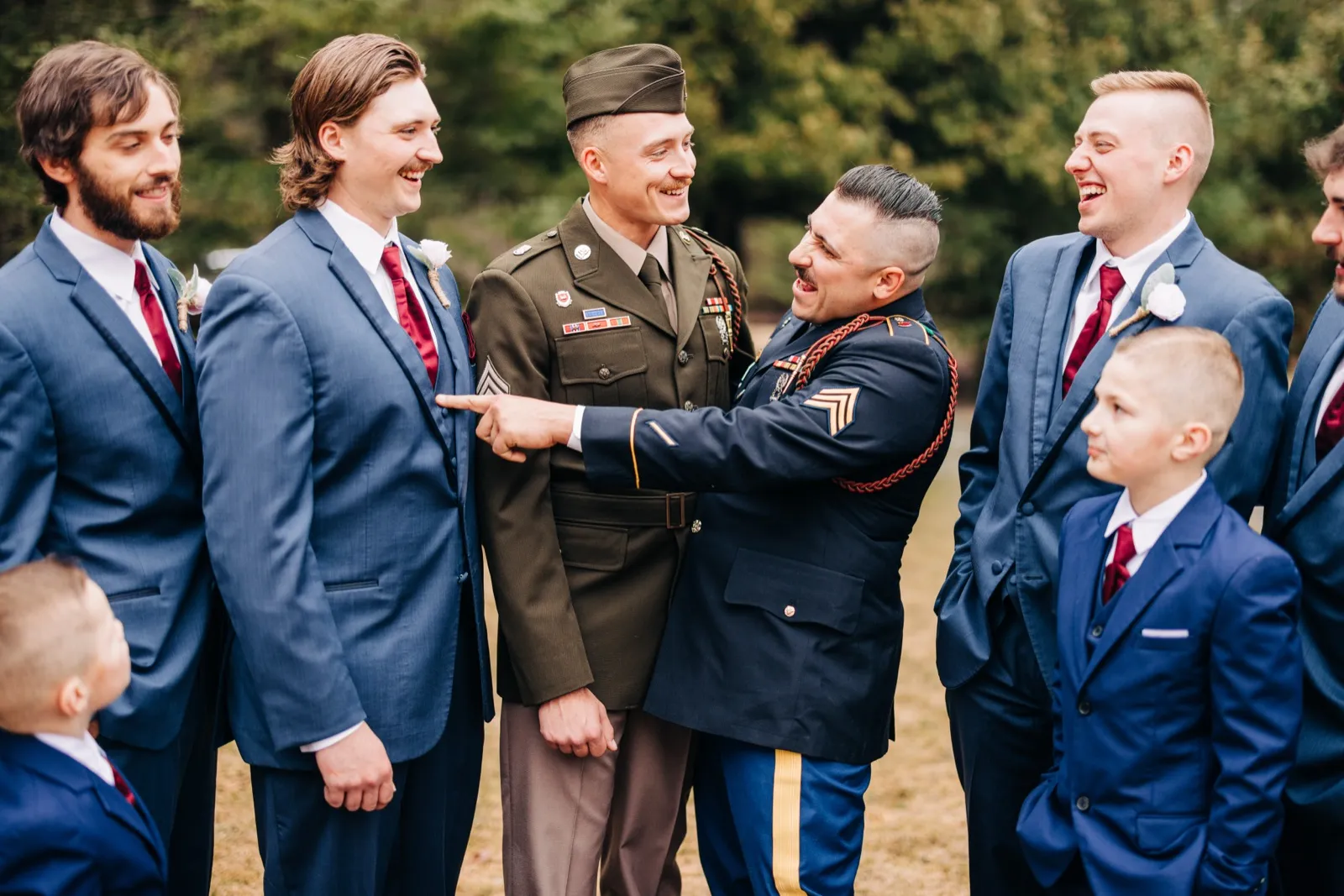 The groom laughs with his groomsmen as a uniformed military guest playfully points at him during an outdoor gathering. — Tim Larsen Photography, Brainerd Lakes MN