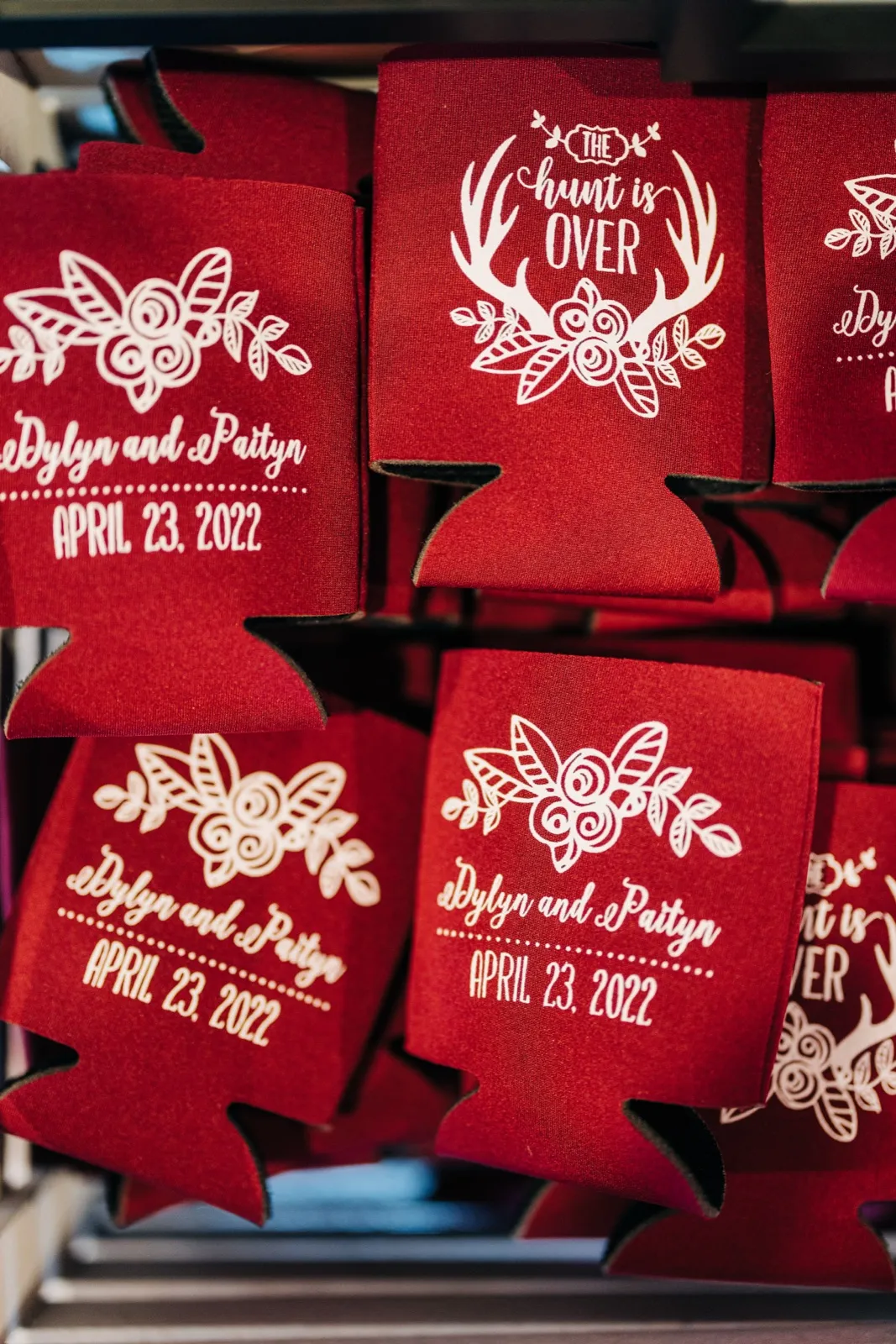 Stacked deep red koozies printed in white with the couple's names, the wedding date April 23, 2022, and a 'The Hunt is Over' antler-and-floral motif. — Tim Larsen Photography, Brainerd Lakes MN