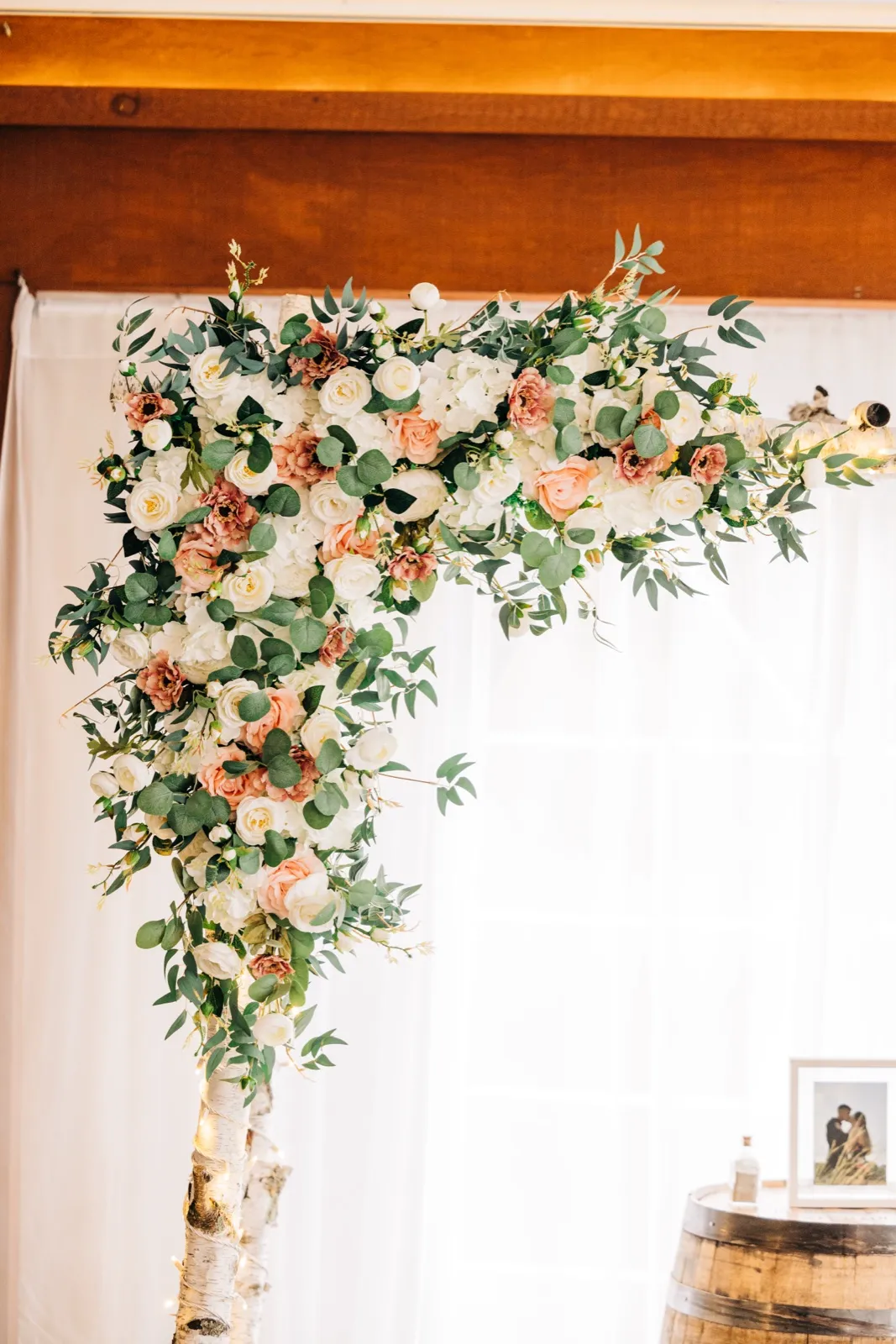 A birch-pole ceremony arch draped with white and blush roses, eucalyptus, and greenery stands in front of a sheer-curtained window with warm wood beams above. — Tim Larsen Photography, Brainerd Lakes MN