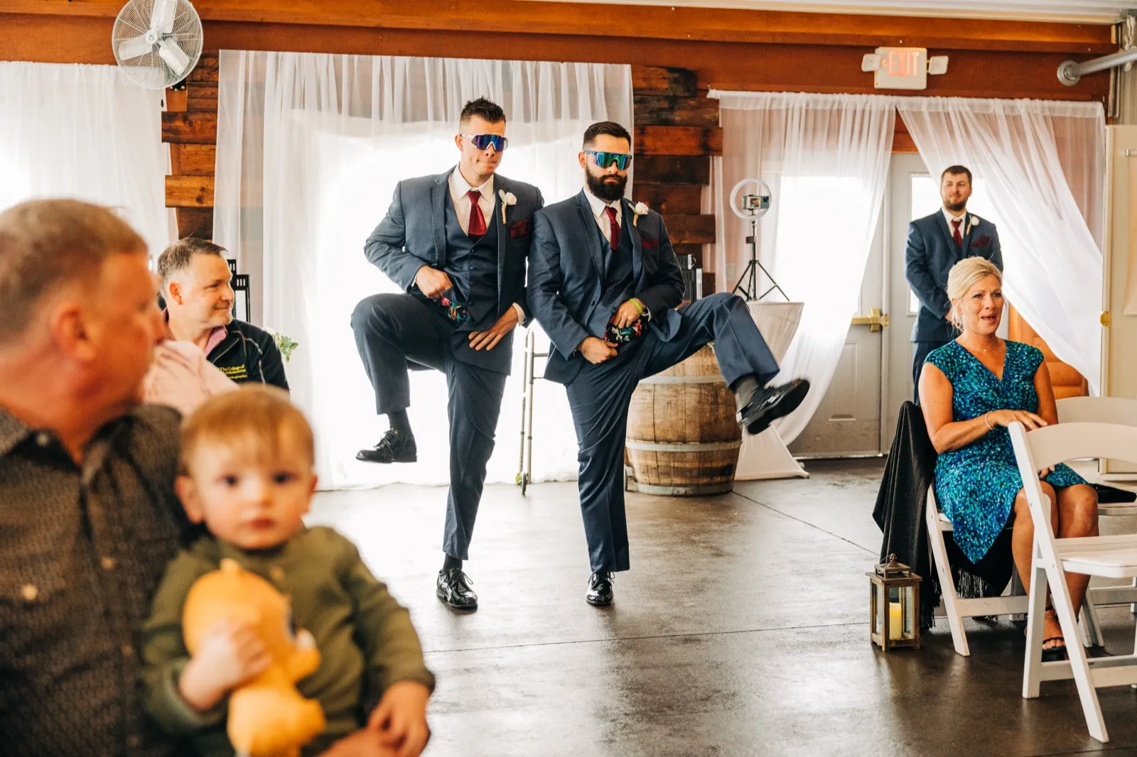Two groomsmen in navy suits and sport sunglasses strike a synchronized leg-kick pose during their reception entrance while seated guests watch. — Tim Larsen Photography, Brainerd Lakes MN