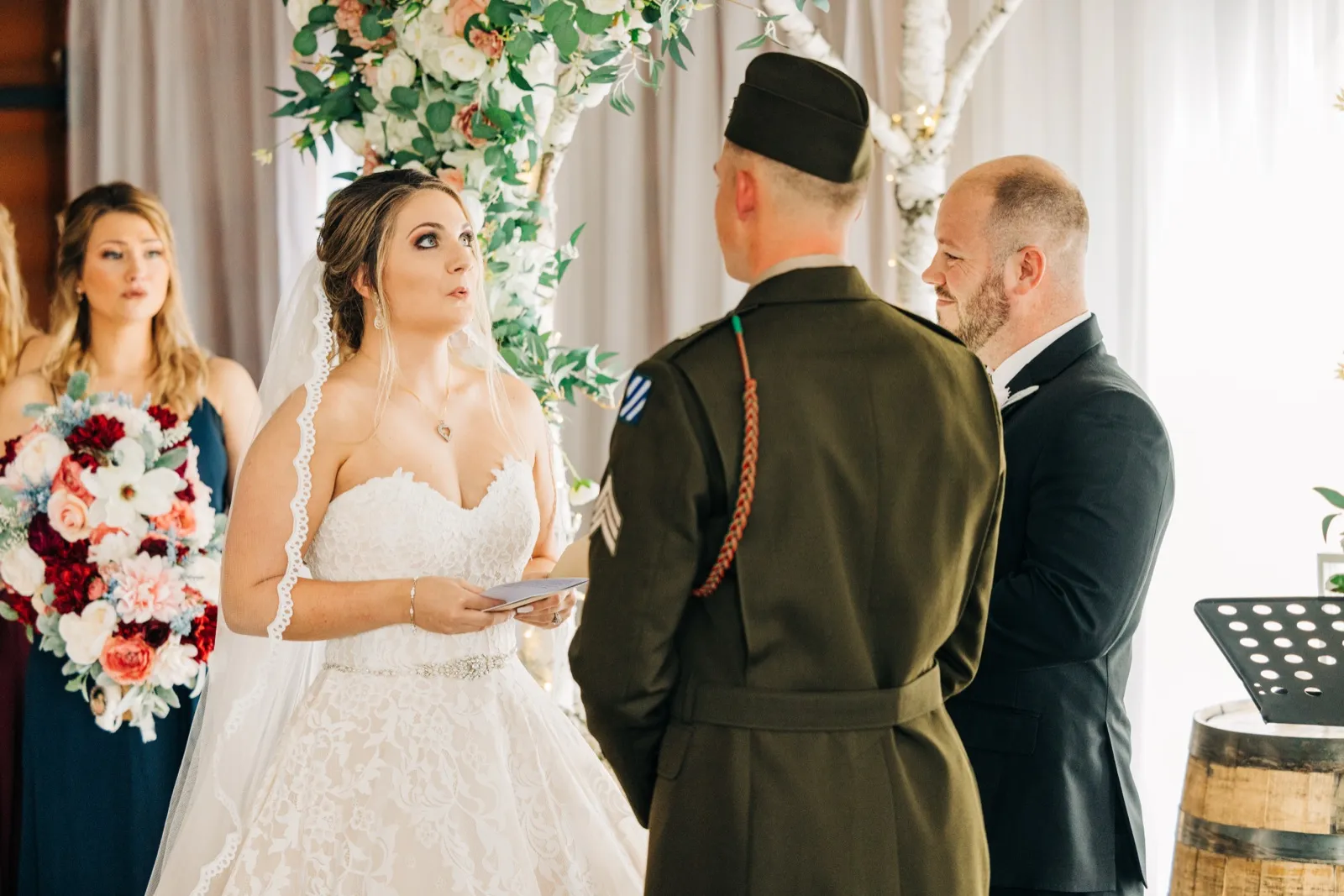 The bride holds her vow card and looks up at her groom, who wears a green military dress uniform, beneath a floral ceremony arch. — Tim Larsen Photography, Brainerd Lakes MN