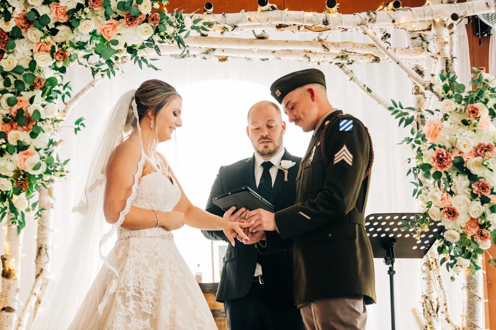 The bride looks down with a soft smile as the groom in dress military uniform places a ring on her finger beneath a birch and floral ceremony arch, with the officiant reading between them. — Tim Larsen Photography, Brainerd Lakes MN