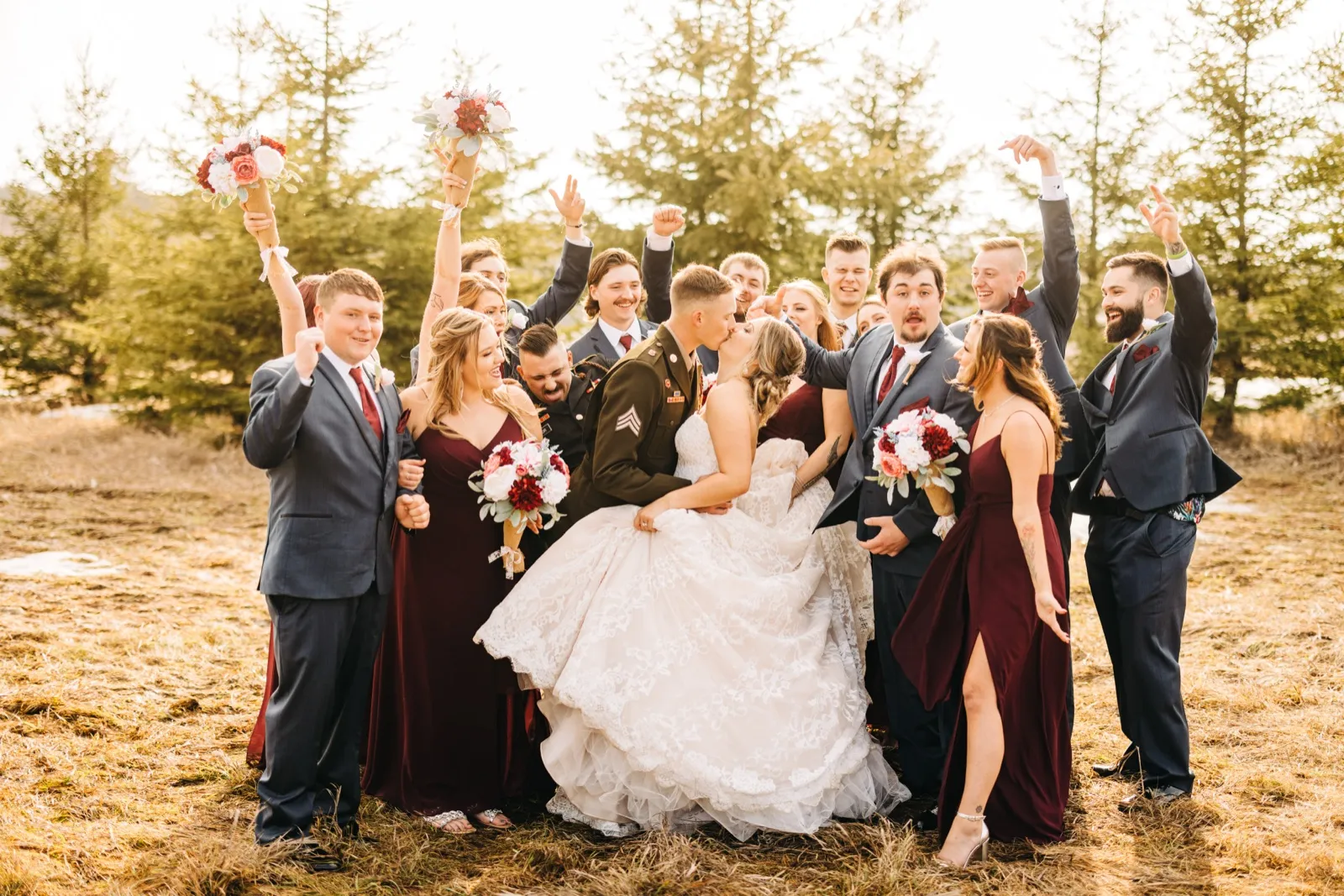 The bride and groom kiss at the center of their cheering bridal party in a sunlit evergreen clearing, with bouquets and arms raised around them. — Tim Larsen Photography, Brainerd Lakes MN