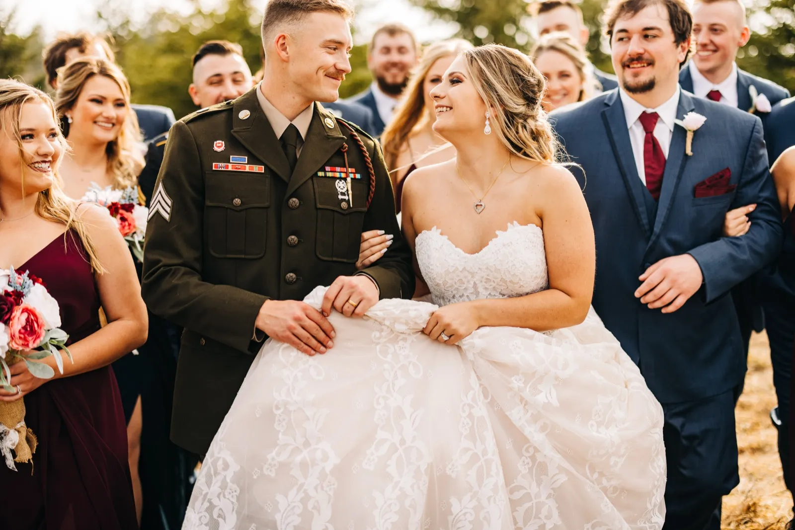 The bride in a strapless lace gown and the groom in an Army dress uniform laugh as they walk surrounded by their bridal party in warm afternoon backlight. — Tim Larsen Photography, Brainerd Lakes MN