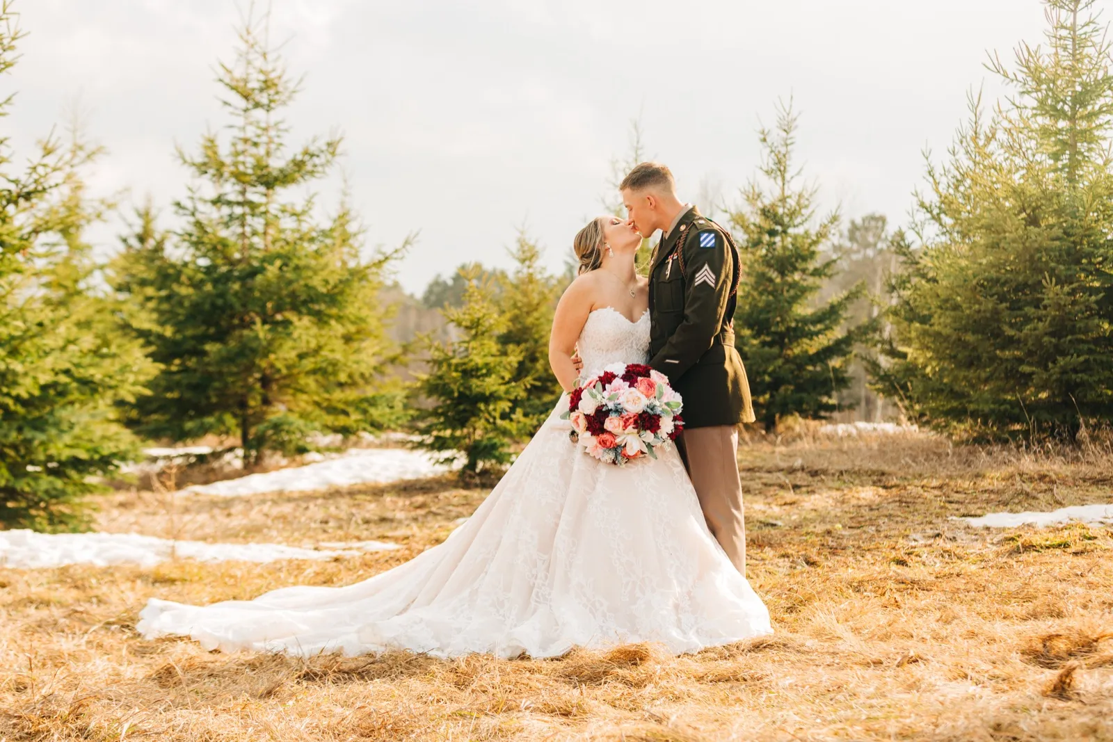 The bride and groom kiss in a clearing surrounded by tall evergreen trees, her gown train fanned across patches of dry grass and lingering snow as he wears his military dress uniform. — Tim Larsen Photography, Brainerd Lakes MN