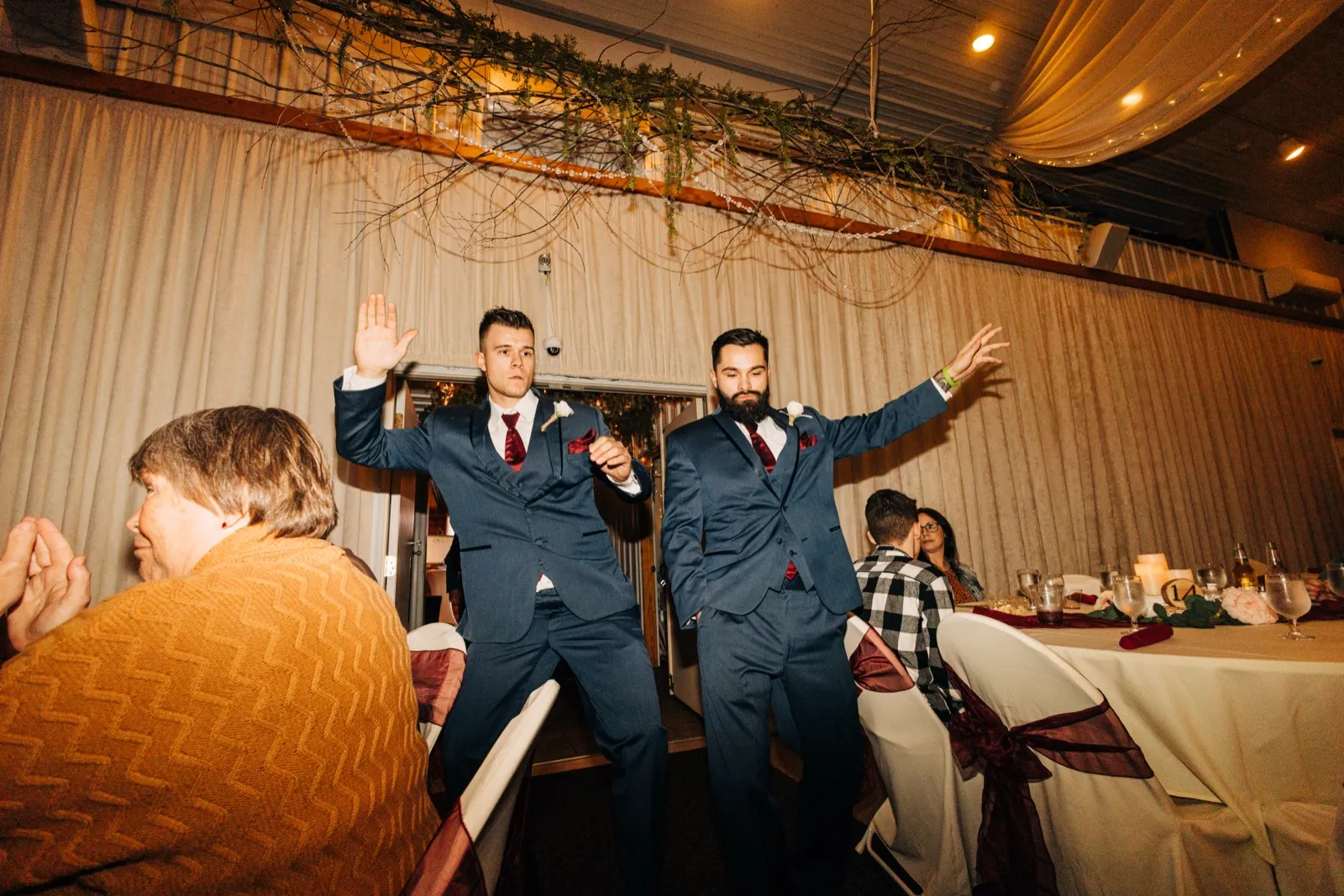 Two groomsmen in navy suits strike exaggerated poses with arms raised as they enter the reception space under draped fabric and greenery. — Tim Larsen Photography, Brainerd Lakes MN