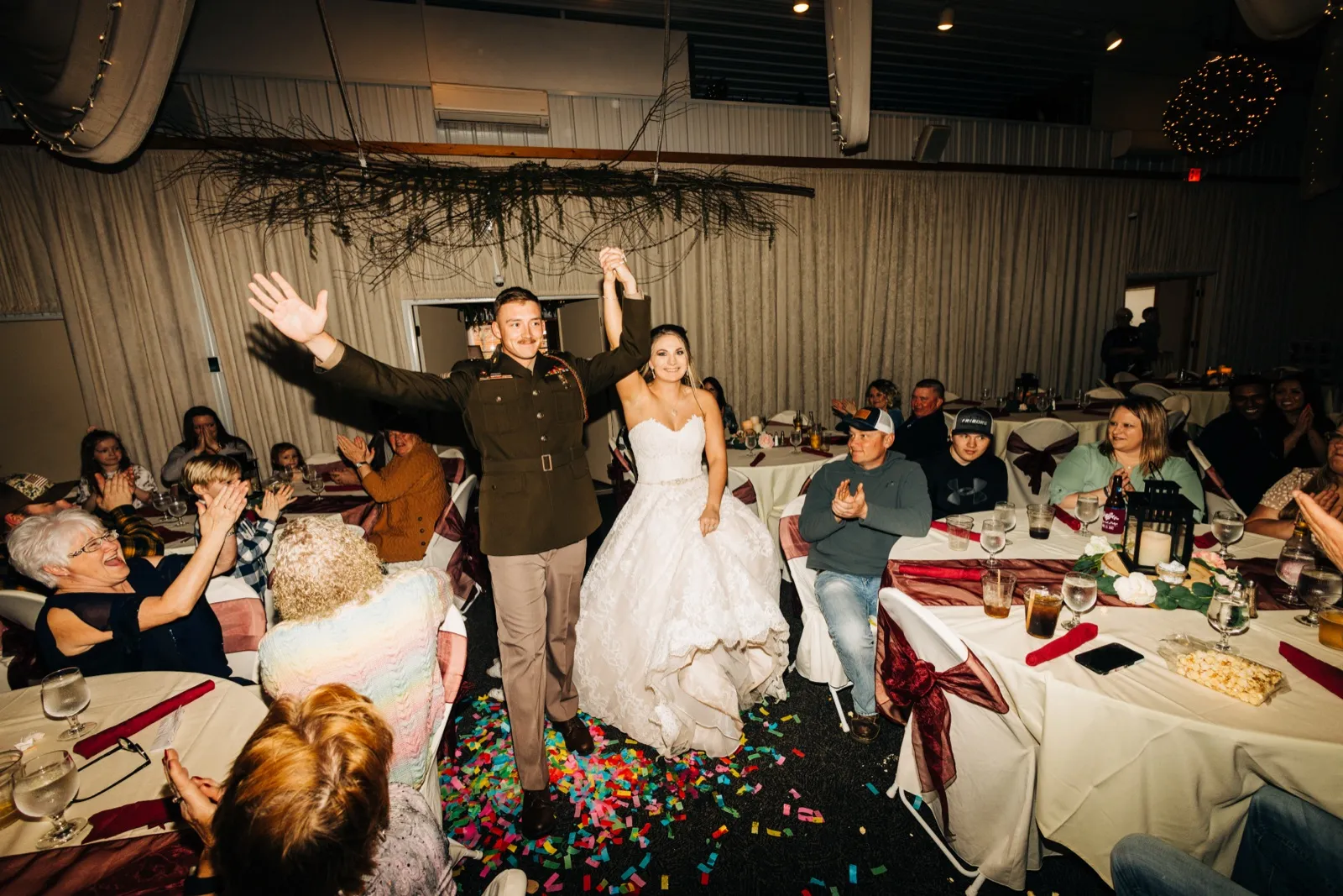 The groom in Marine dress uniform and the bride in a white ballgown raise their joined hands triumphantly as they enter their reception, surrounded by clapping guests and confetti scattered across the floor. — Tim Larsen Photography, Brainerd Lakes MN