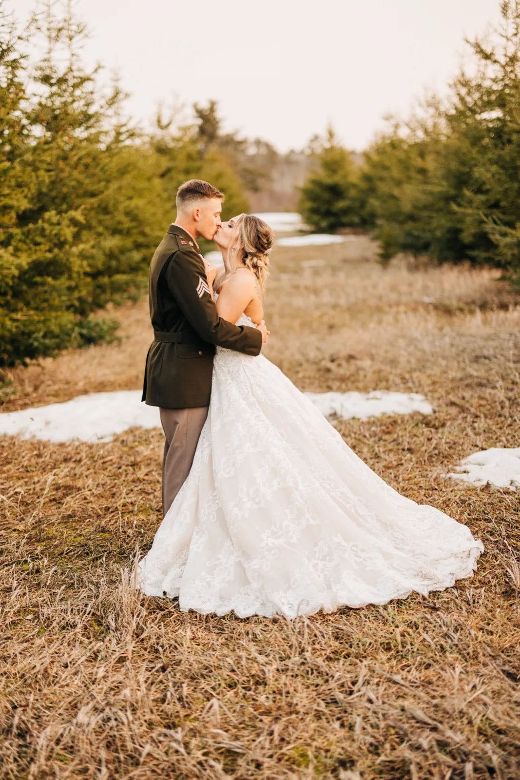 A bride in a long lace ballgown kisses her groom in a military dress uniform in a clearing flanked by tall evergreen trees with patches of snow on the ground. — Tim Larsen Photography, Brainerd Lakes MN