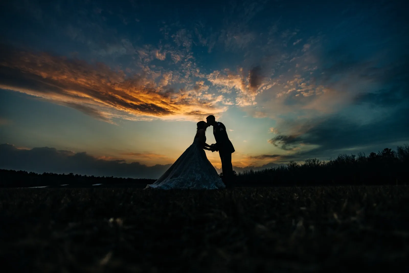 The bride and groom kiss in silhouette beneath a vast orange and blue sunset sky, the bride's full gown trailing across an open field. — Tim Larsen Photography, Brainerd Lakes MN