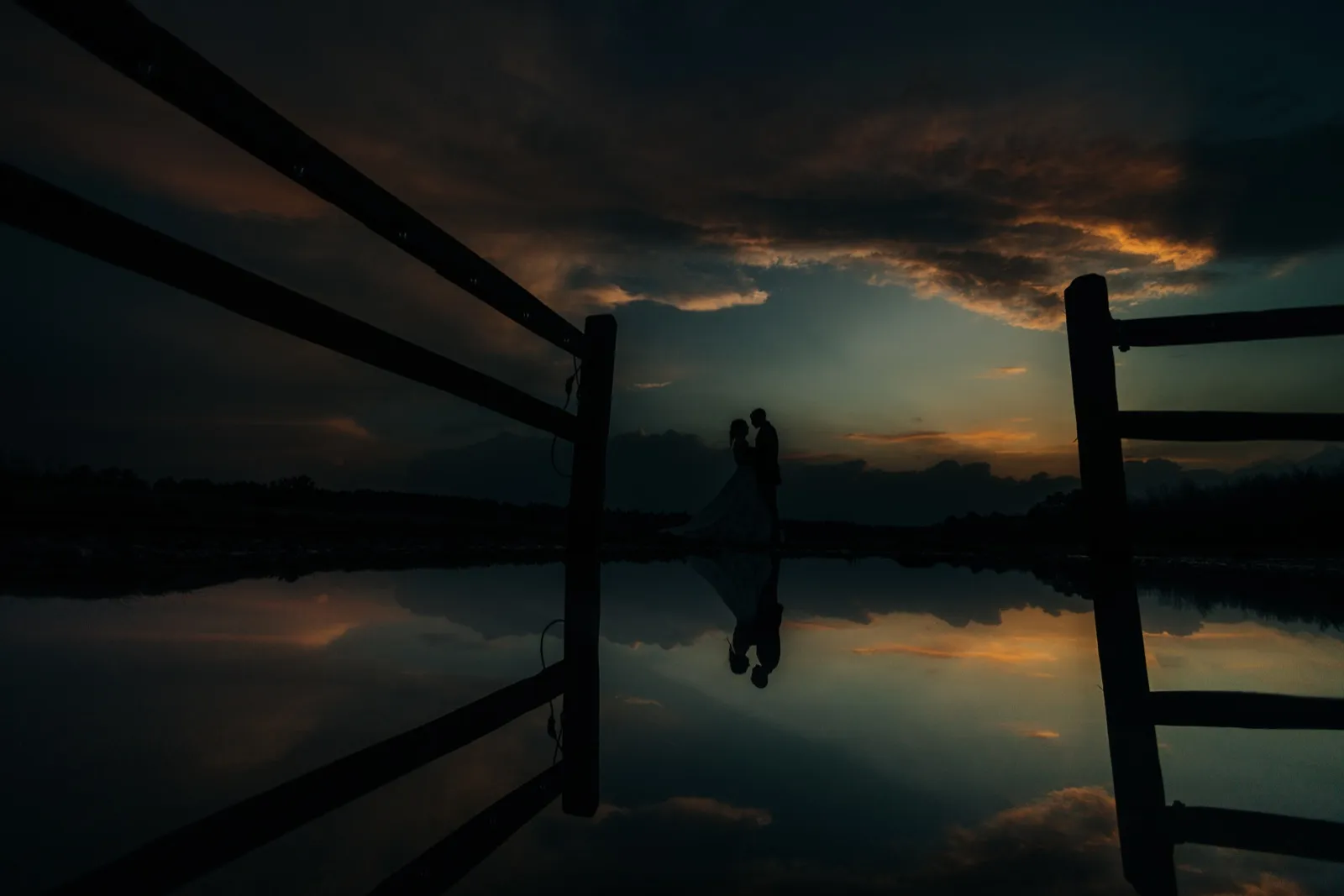 The couple stands in silhouette against a dusk sky, framed between wooden fence rails, with their reflection mirrored in the still pond water below. — Tim Larsen Photography, Brainerd Lakes MN