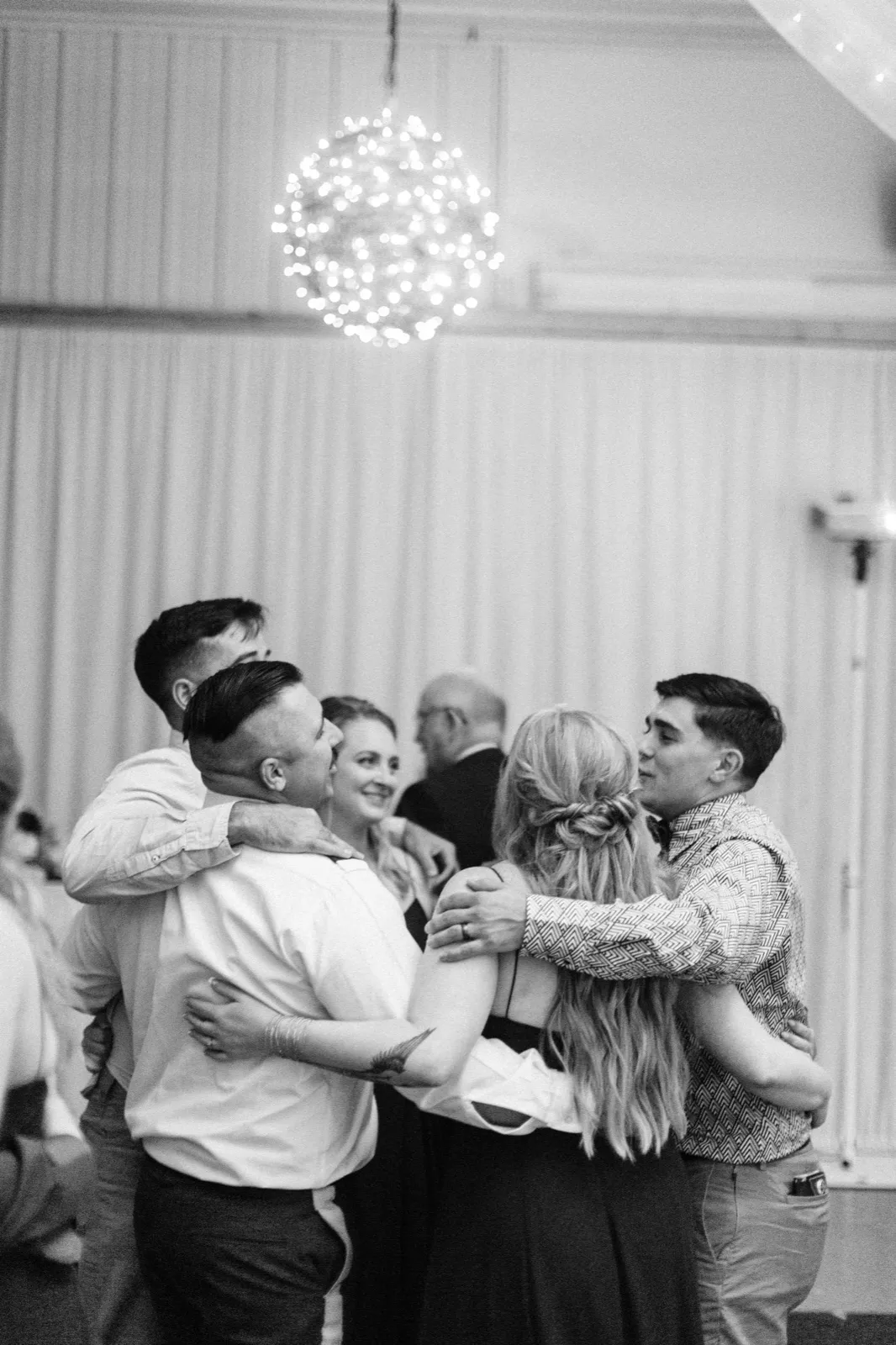 A group of guests embraces in a circle on the dance floor beneath a glowing sphere chandelier, photographed in black and white. — Tim Larsen Photography, Brainerd Lakes MN