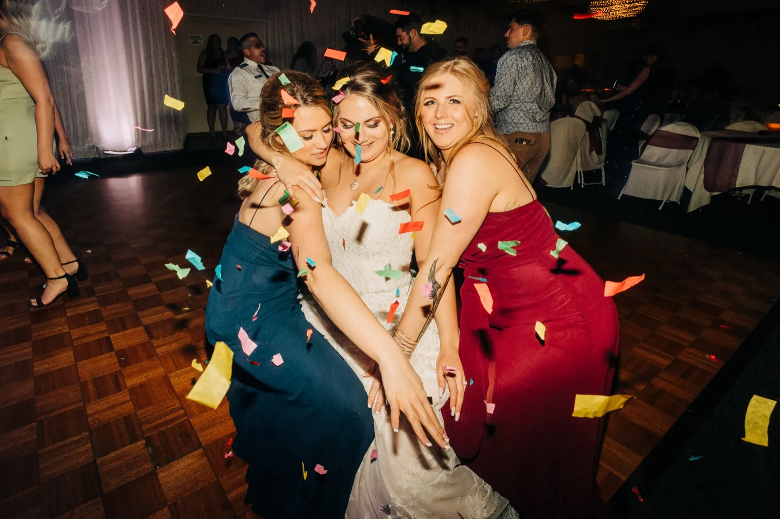 The bride and two bridesmaids in navy and burgundy dresses embrace on the dance floor as colorful confetti falls around them. — Tim Larsen Photography, Brainerd Lakes MN
