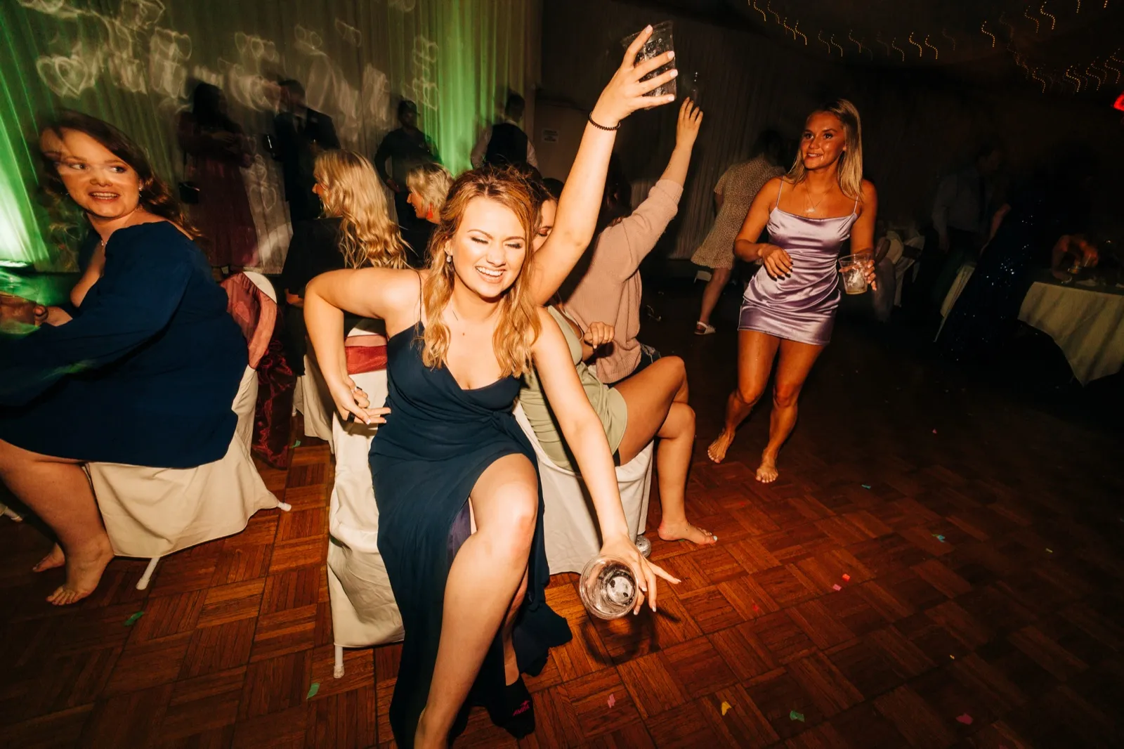 A guest in a navy dress dances mid-motion on a wooden dance floor, holding a glass low, while other guests dance around her under green stage lighting. — Tim Larsen Photography, Brainerd Lakes MN