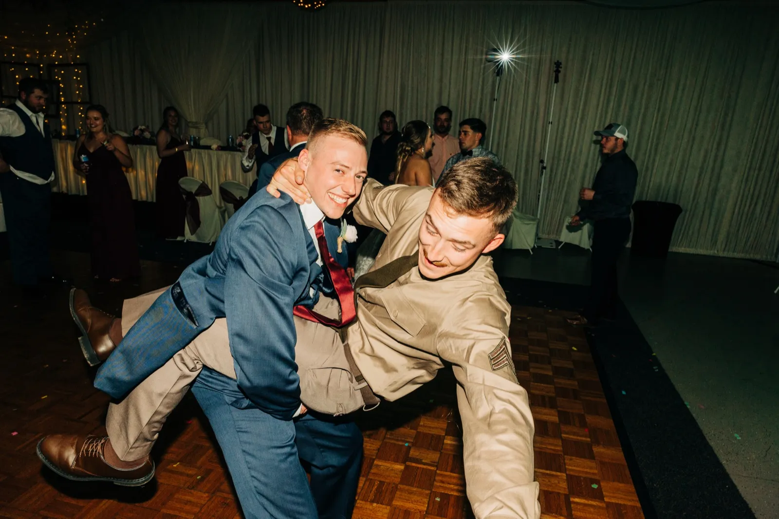 The groom grins as he wrestles playfully with a guest in military dress on the reception dance floor, both men leaning into each other under flash light. — Tim Larsen Photography, Brainerd Lakes MN