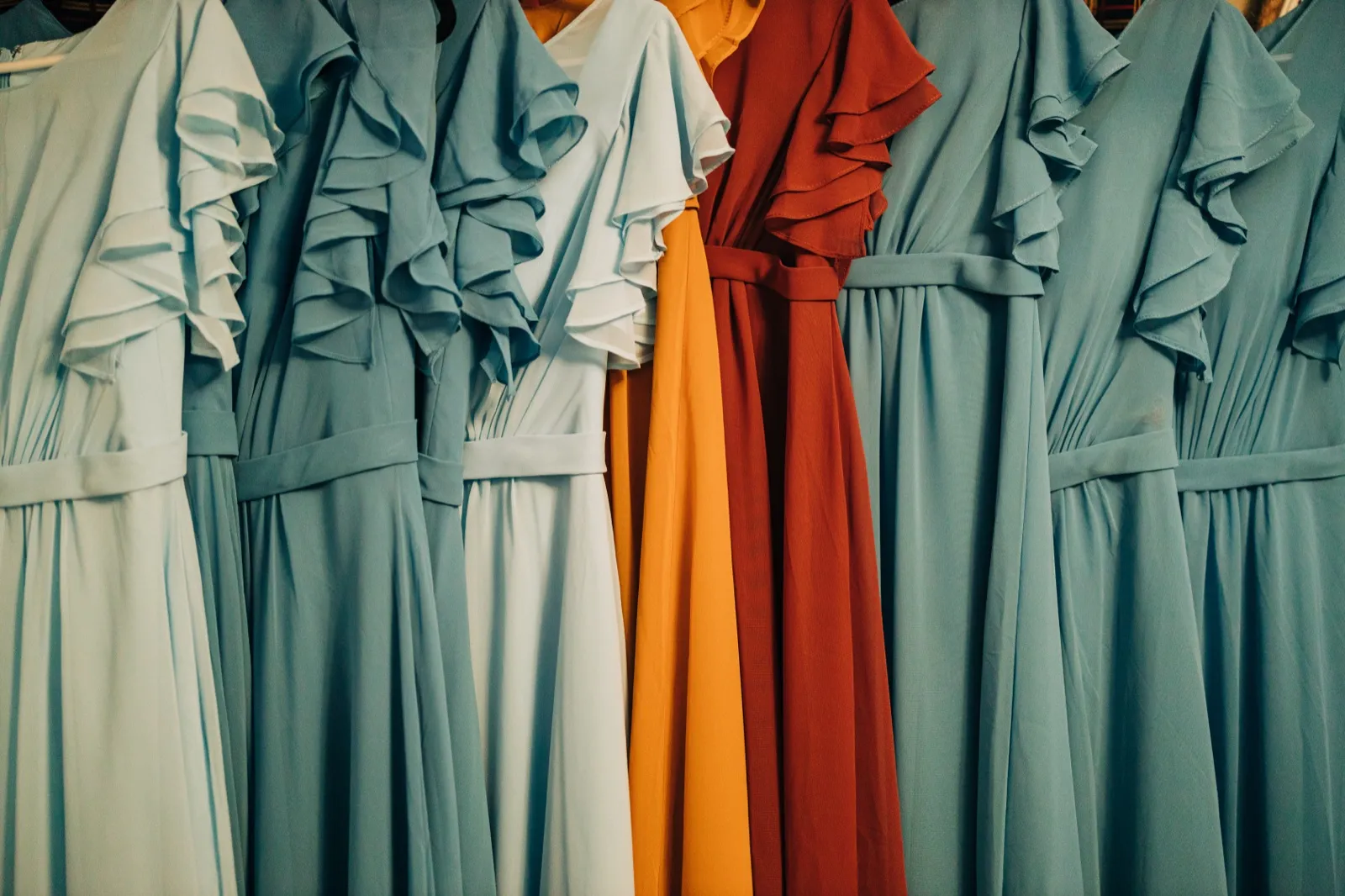 A row of bridesmaid dresses hangs side by side, with dusty blue gowns flanking a rust-red and a marigold dress in the center. — Tim Larsen Photography, Brainerd Lakes MN