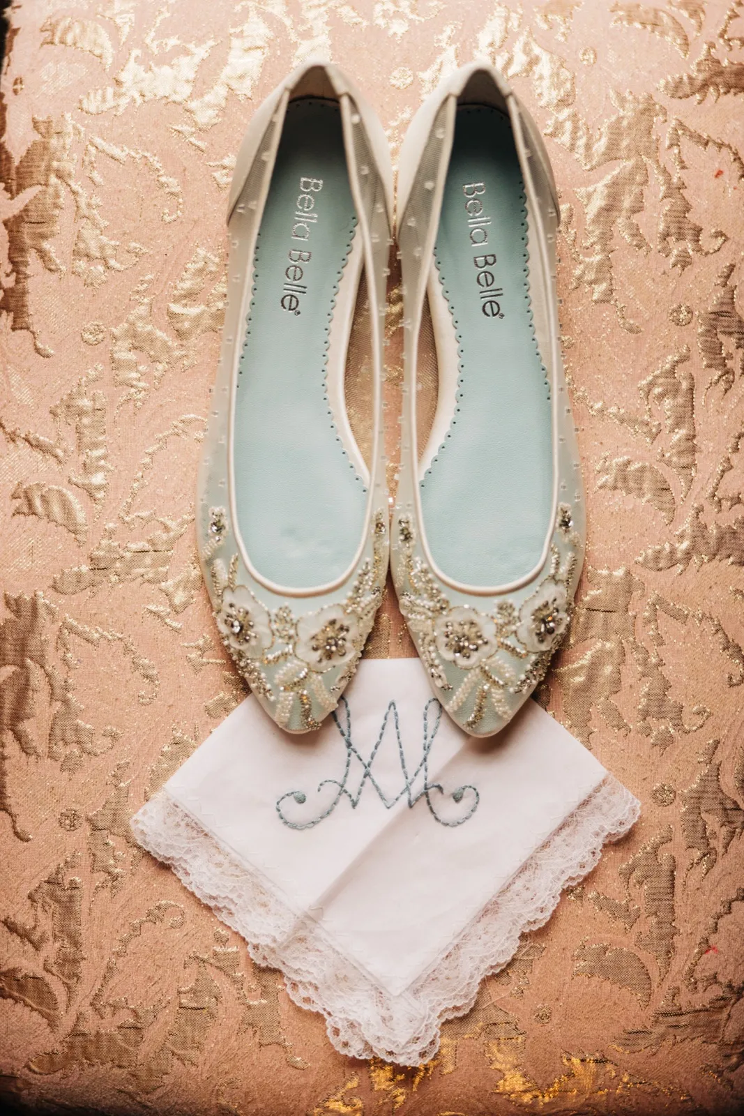 A pair of beaded ivory Bella Belle bridal flats sits above an embroidered monogrammed lace handkerchief on a gold brocade surface. — Tim Larsen Photography, Brainerd Lakes MN