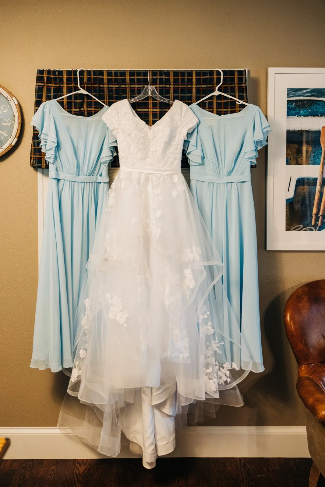 A white lace wedding gown hangs centered between two dusty-blue bridesmaid dresses against a plaid backdrop on a house wall. — Tim Larsen Photography, Brainerd Lakes MN