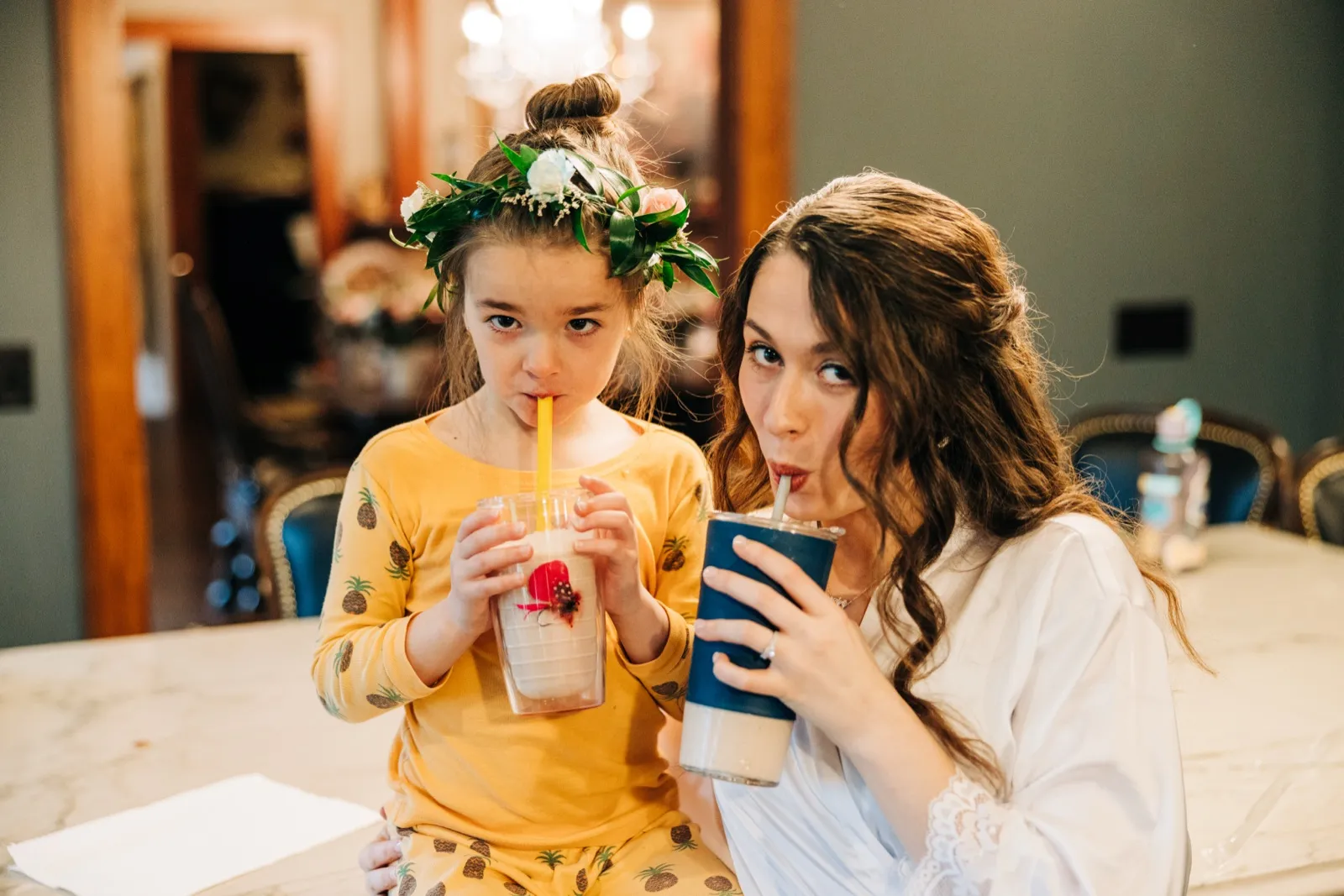 The bride in a white robe and a young flower girl in pineapple pajamas with a flower crown sip from straws side by side, both looking toward the camera. — Tim Larsen Photography, Brainerd Lakes MN