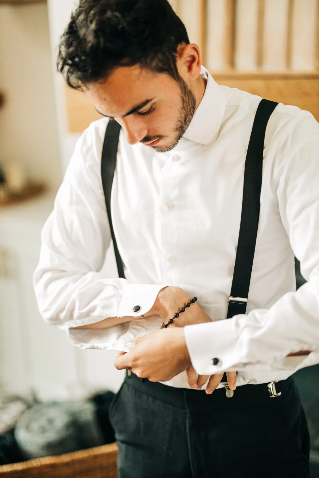 The groom looks down as he fastens a cufflink, wearing a white dress shirt and black suspenders, with a beaded bracelet on his wrist. — Tim Larsen Photography, Brainerd Lakes MN