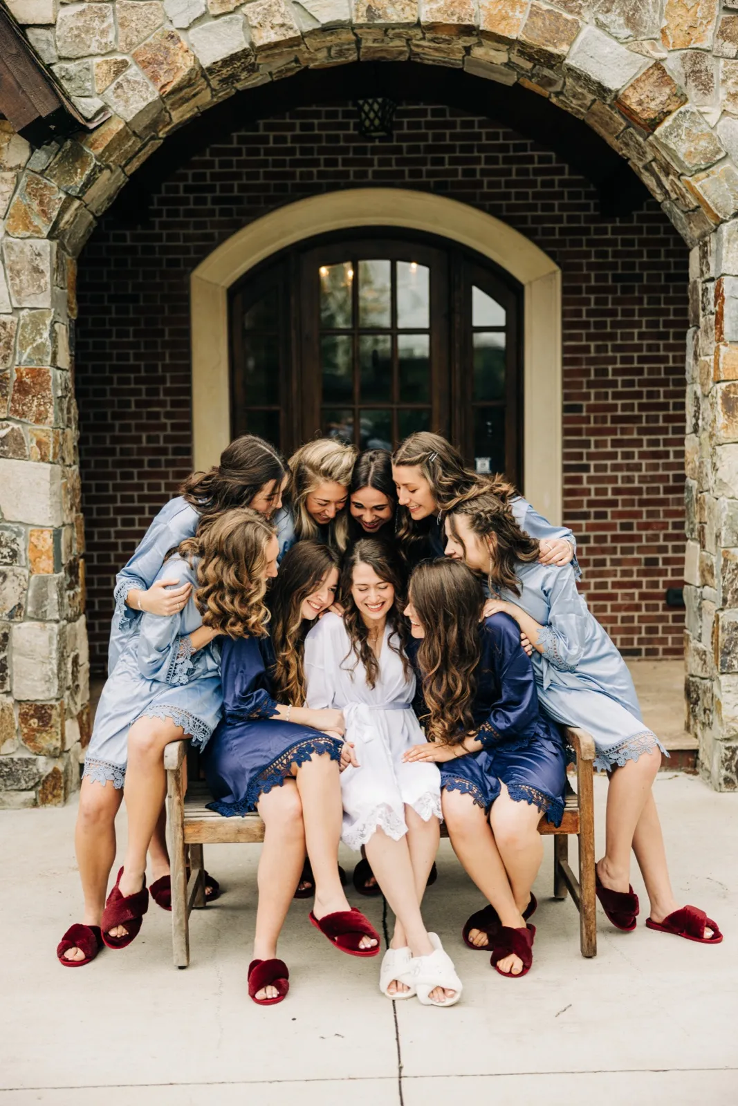 The bride in a white robe sits centered on a wooden bench, surrounded by seven bridesmaids in matching dusty-blue robes leaning in to embrace her beneath a stone archway. — Tim Larsen Photography, Brainerd Lakes MN