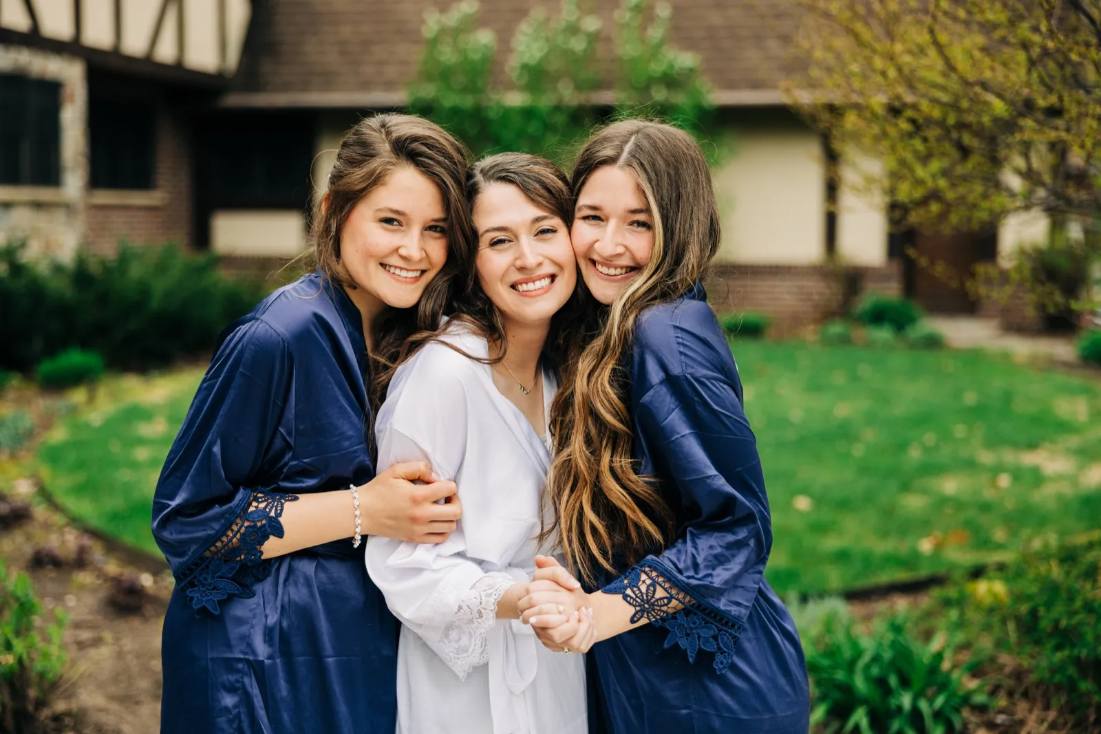 The bride in a white robe is pressed cheek-to-cheek between two bridesmaids in navy lace-trimmed robes, all smiling in front of a Tudor-style house. — Tim Larsen Photography, Brainerd Lakes MN