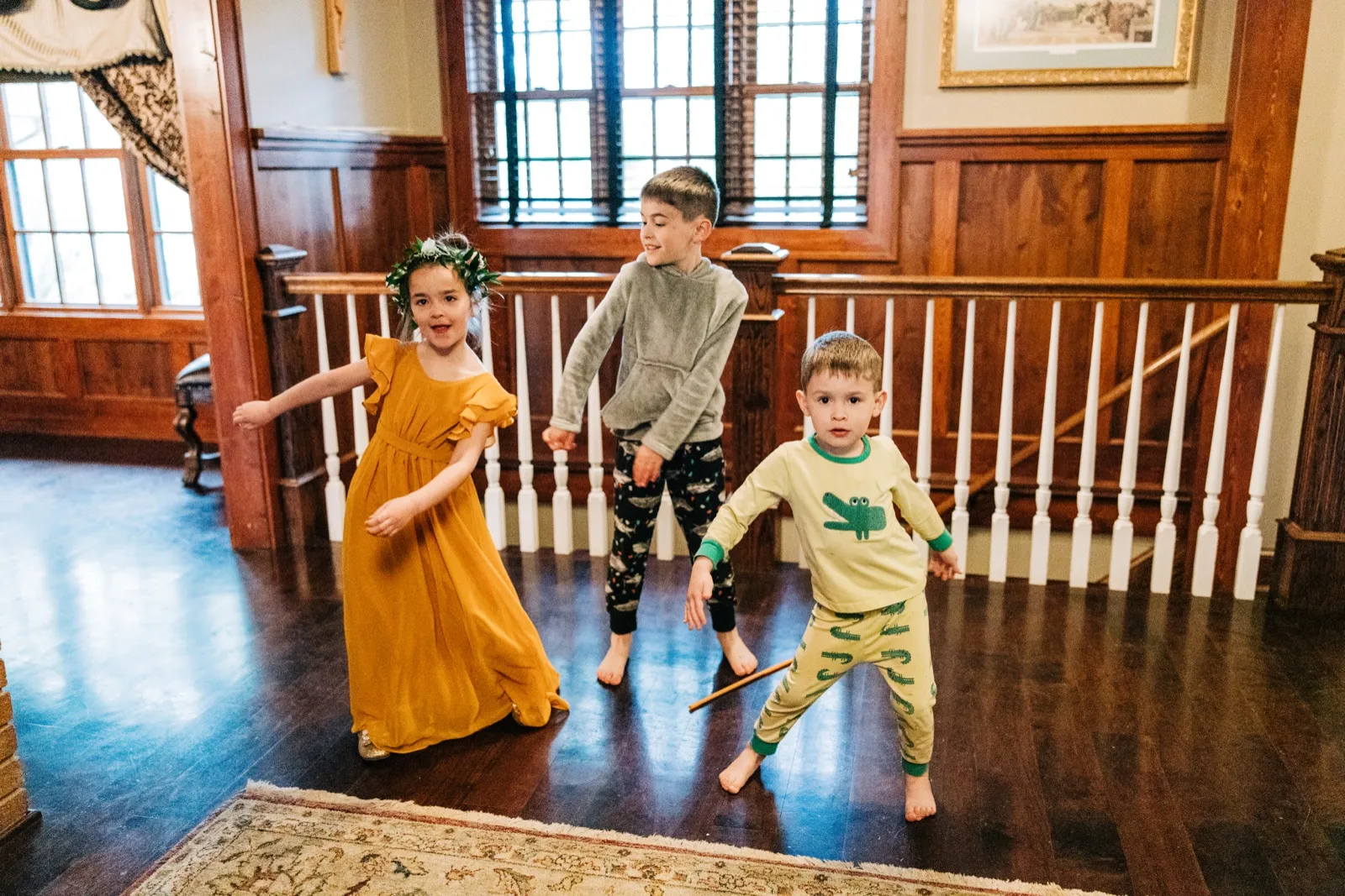 Three young children dance in a wood-paneled upstairs hallway, the flower girl in a mustard dress and floral crown beside two boys in pajamas. — Tim Larsen Photography, Brainerd Lakes MN
