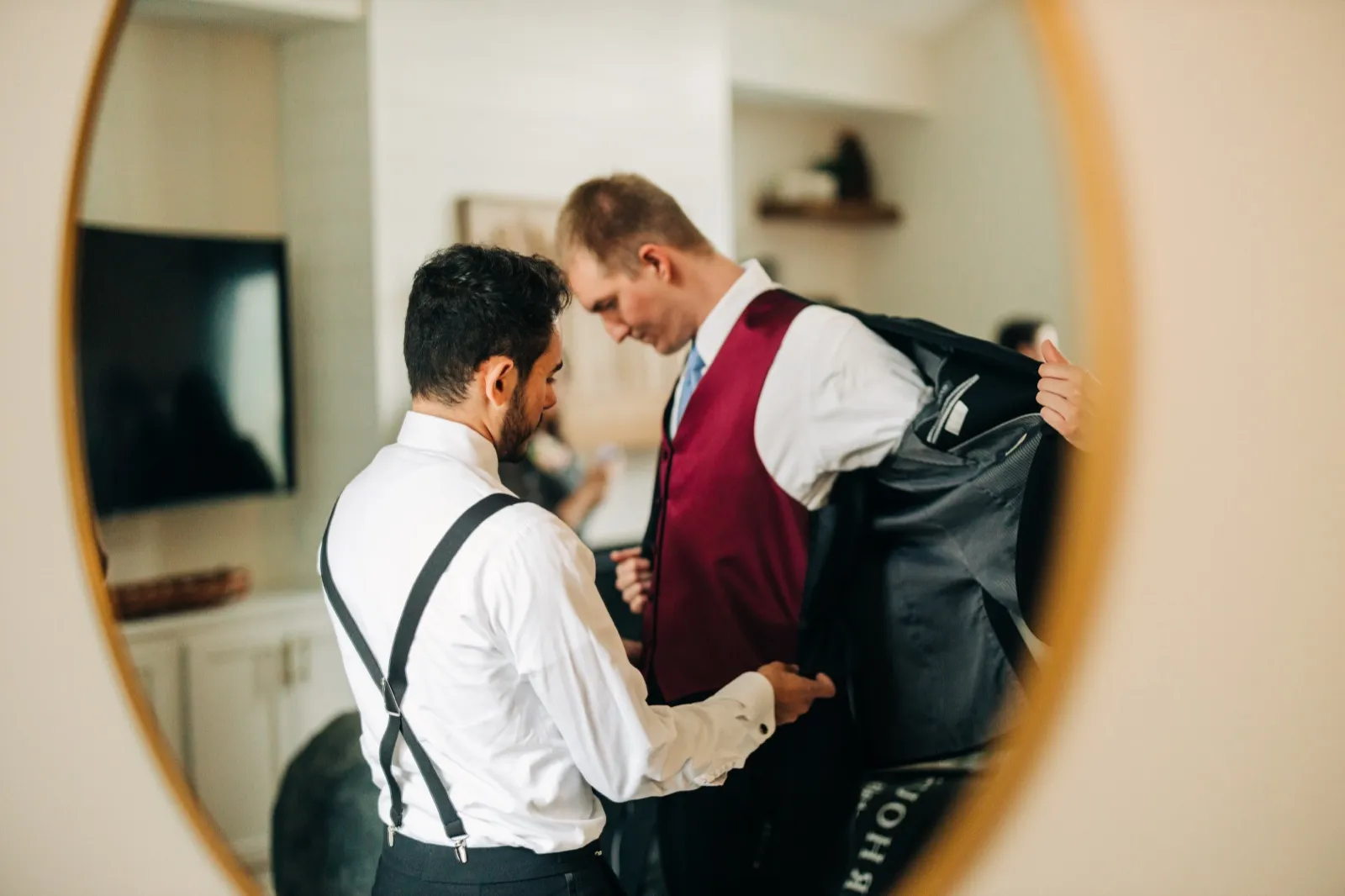 A groom in a burgundy vest holds open his jacket while a groomsman in suspenders helps him, the entire scene reflected in a round wall mirror. — Tim Larsen Photography, Brainerd Lakes MN