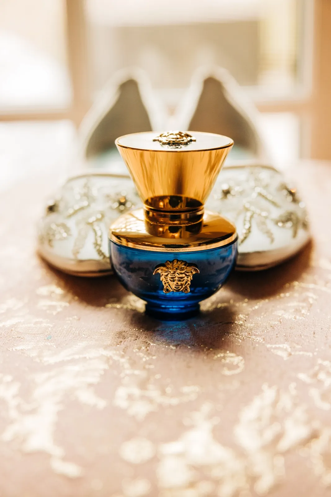 A blue and gold Versace perfume bottle sits centered in front of softly blurred embellished bridal shoes on a textured pink linen. — Tim Larsen Photography, Brainerd Lakes MN