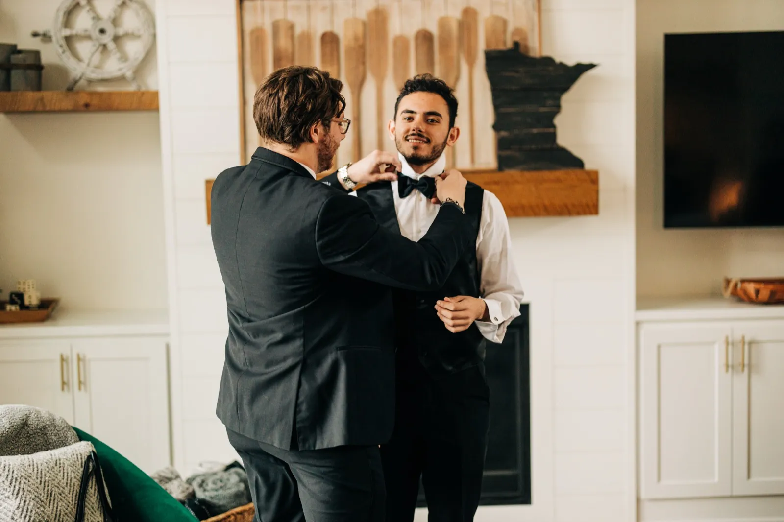 A groomsman in a dark suit adjusts the groom's black bow tie inside a house living room with a wooden Minnesota cutout and oar decor on the wall. — Tim Larsen Photography, Brainerd Lakes MN