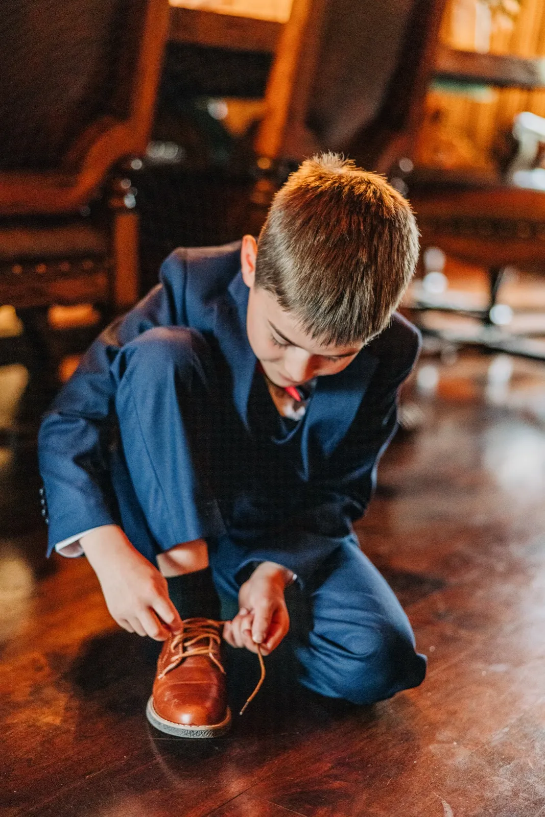 A young boy in a navy suit kneels on a polished wood floor to tie the laces of his brown dress shoe. — Tim Larsen Photography, Brainerd Lakes MN