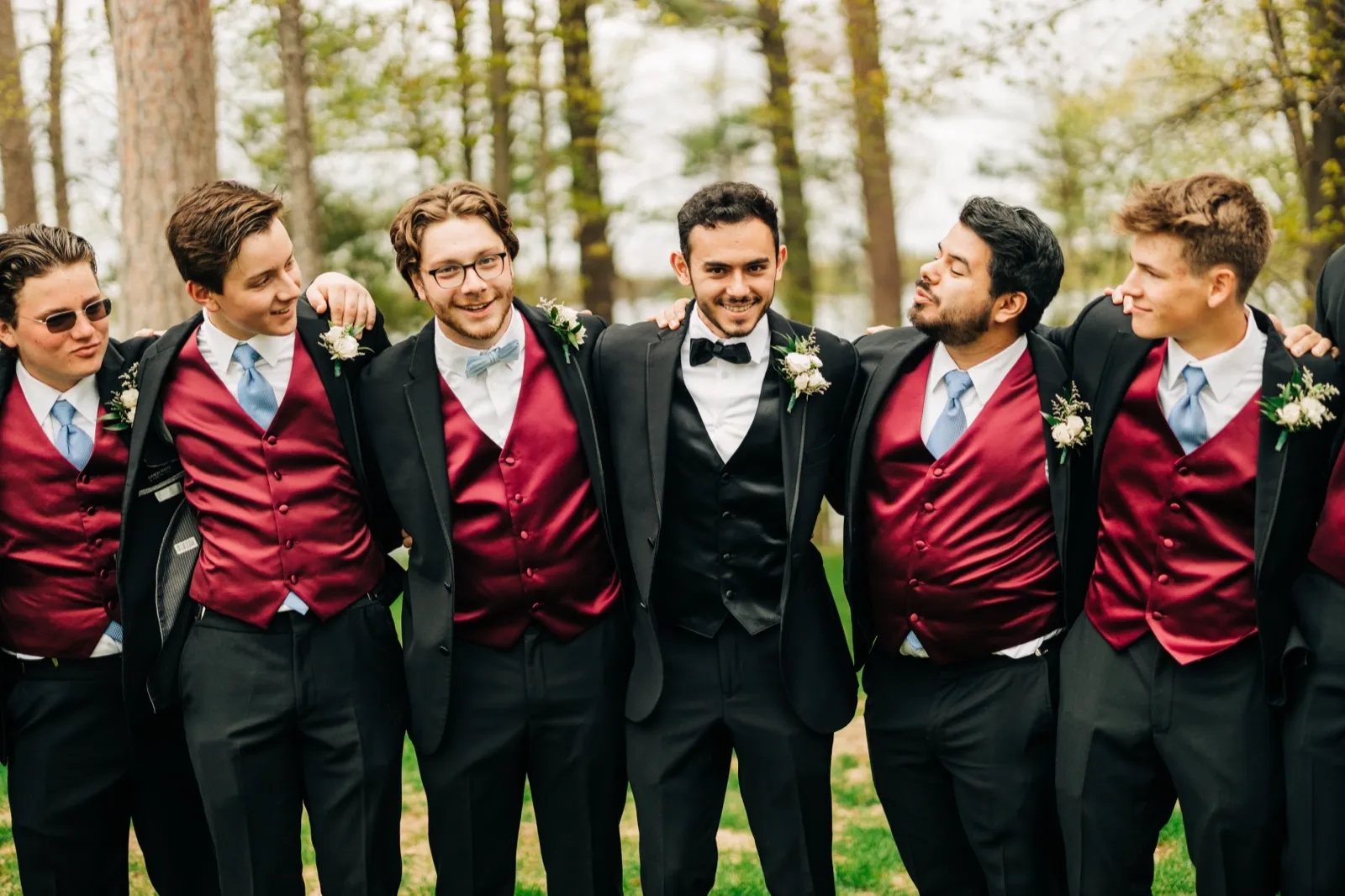 Six groomsmen in burgundy vests and dusty blue ties stand shoulder to shoulder on a lawn, several making playful faces while the groom smiles at the camera. — Tim Larsen Photography, Brainerd Lakes MN