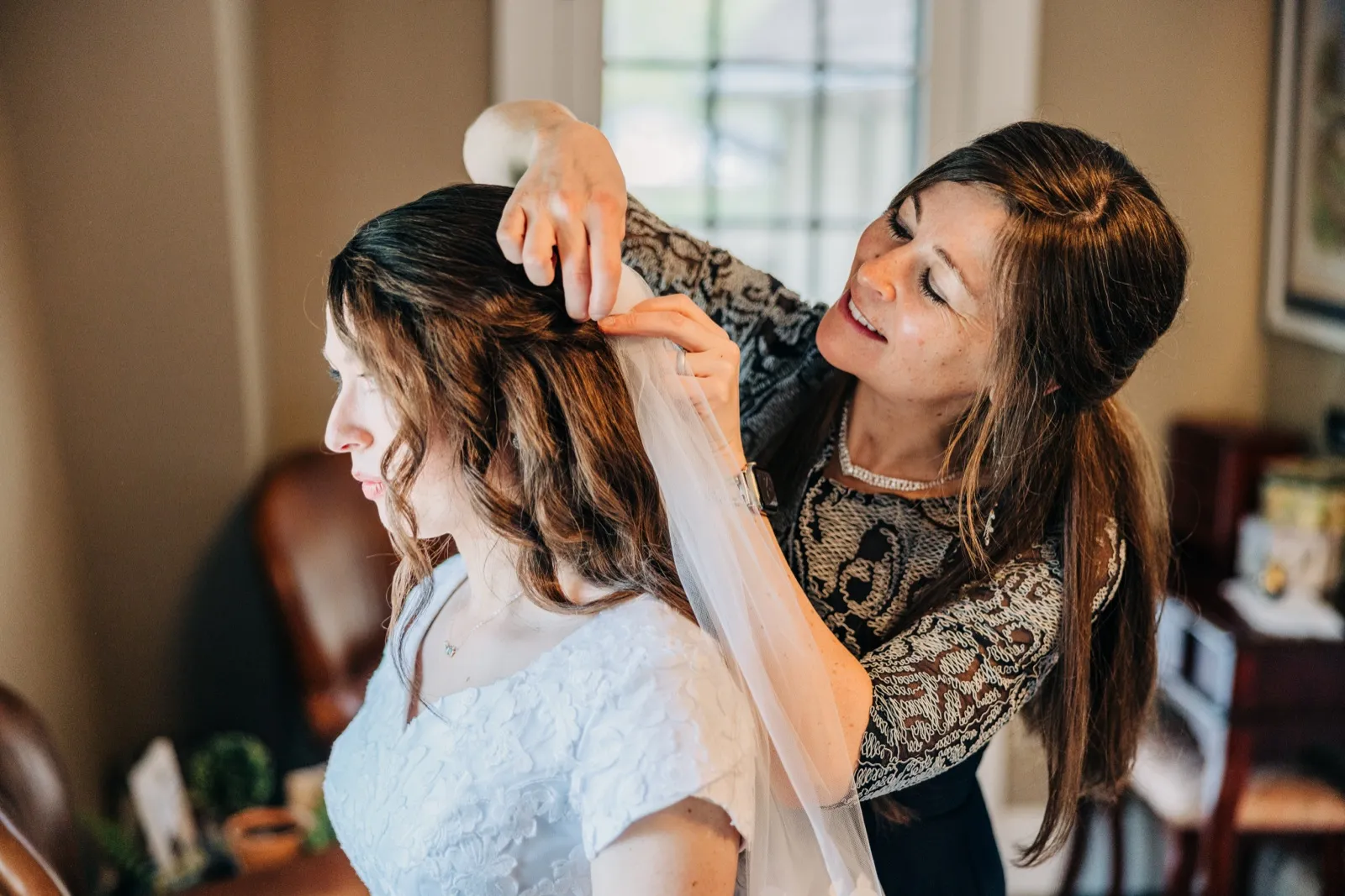 A woman in a lace top fastens a veil into the bride's hair as the bride sits quietly in window light. — Tim Larsen Photography, Brainerd Lakes MN
