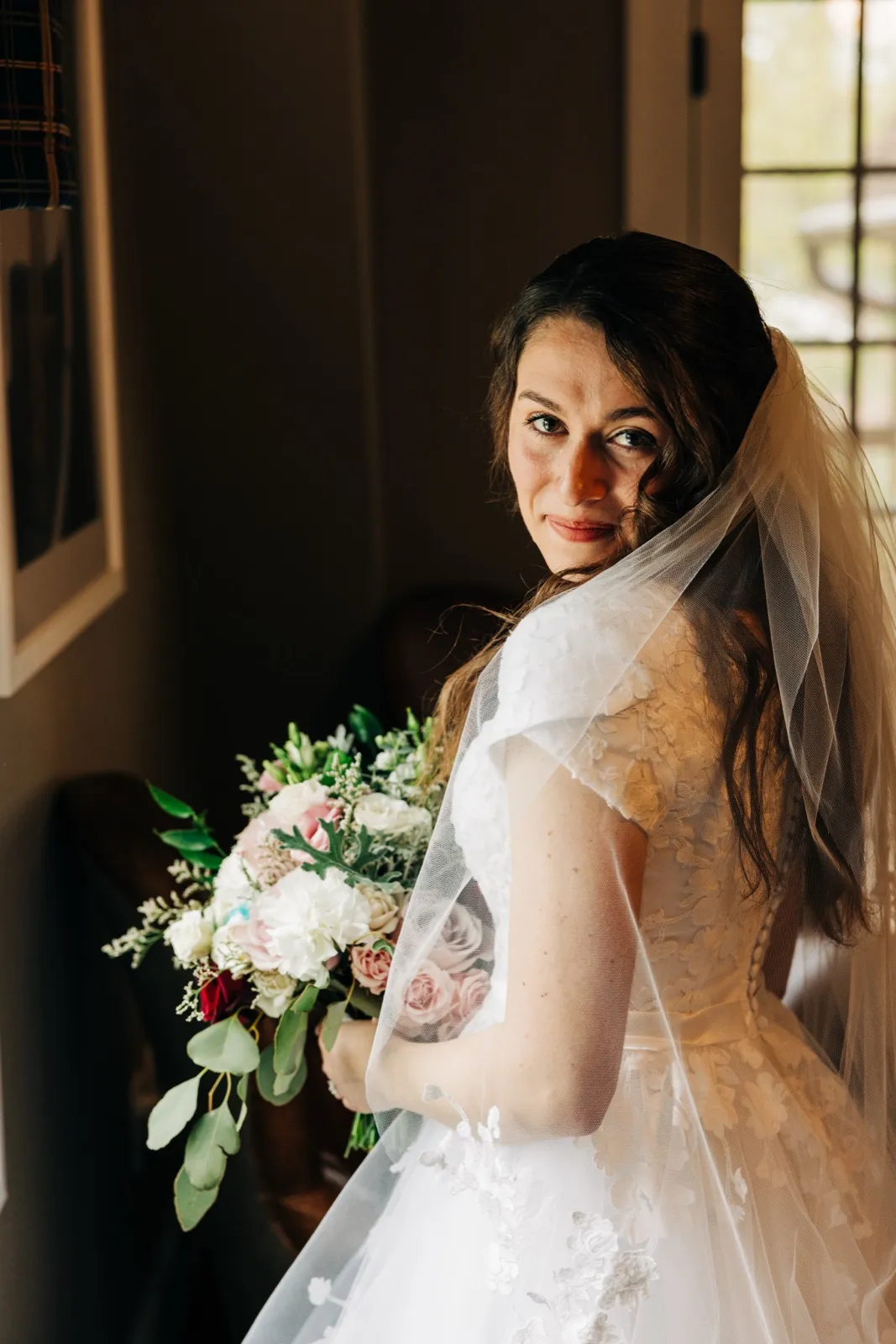 The bride looks over her shoulder toward the camera, her veil draped across her lace gown as she holds a bouquet of pink, white, and burgundy florals near a sunlit house window. — Tim Larsen Photography, Brainerd Lakes MN